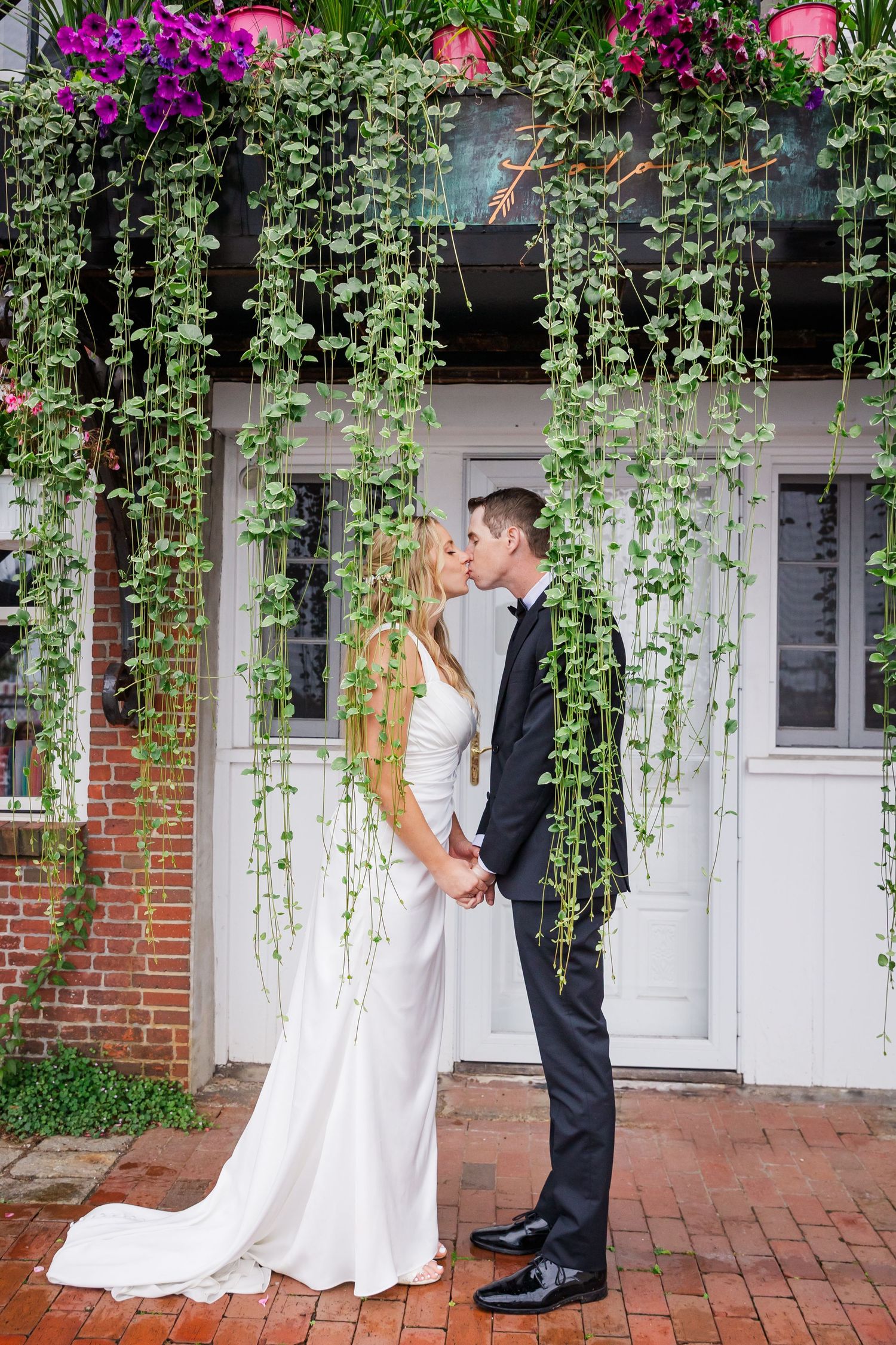 Wedding couple shares intimate moment against white wall with cascading vines and pink flowers.