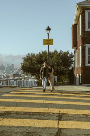 A couple walks together on a yellow striped crosswalk with a street lamp and urban buildings in the background.