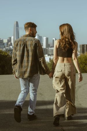 Couple walking hand in hand away from camera with city skyline in background.
