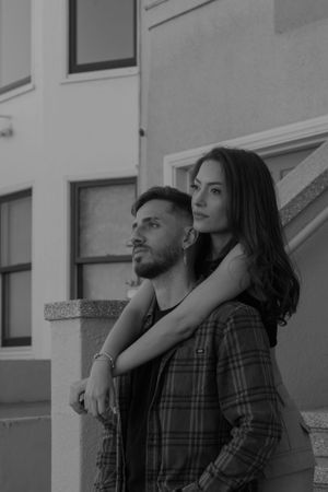 Black and white portrait of couple in casual attire leaning against building exterior.