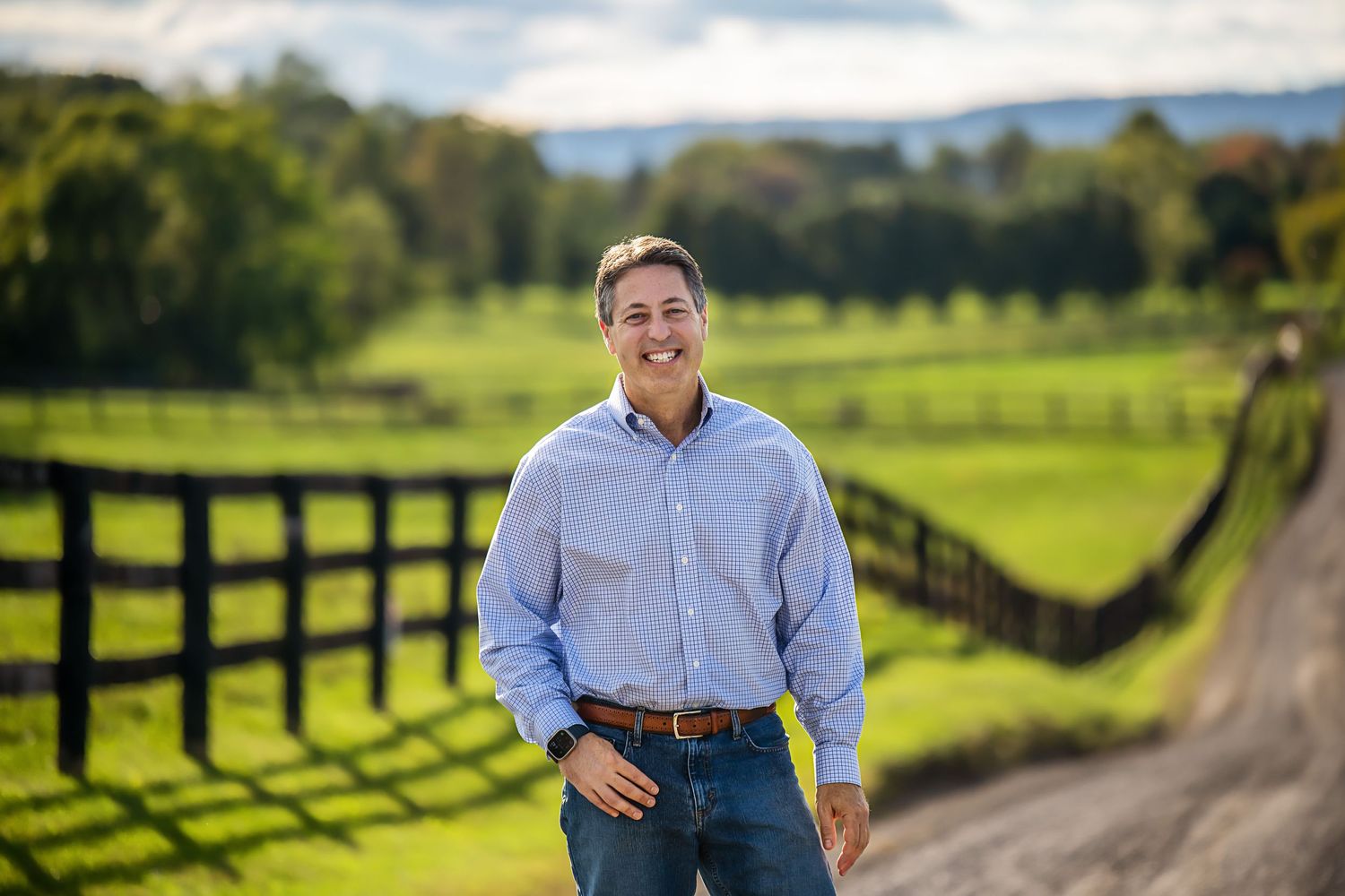 Person in light blue button-down shirt stands by wooden fence on rural country road.