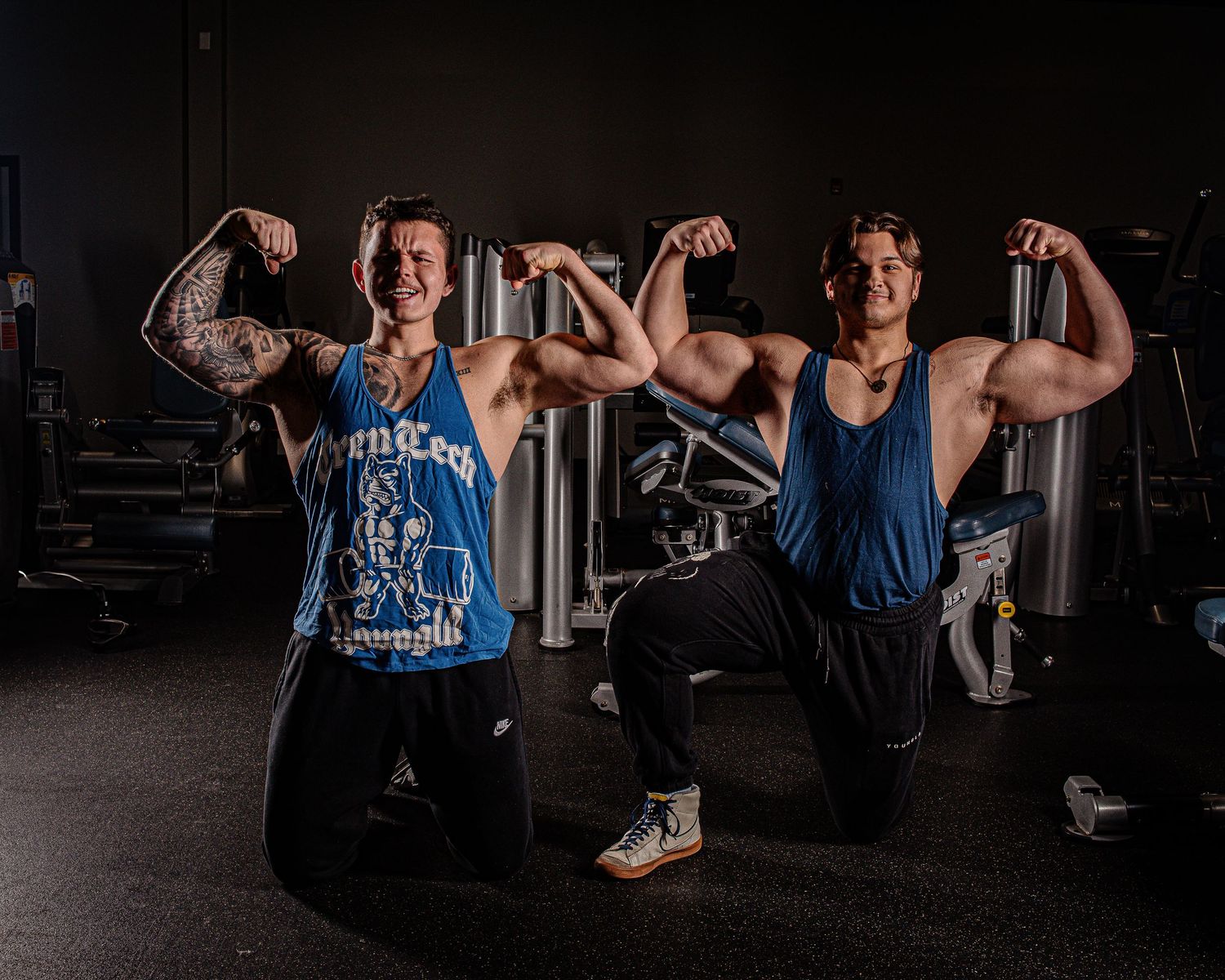 Two bodybuilders flexing muscles in blue tank tops at a gym.