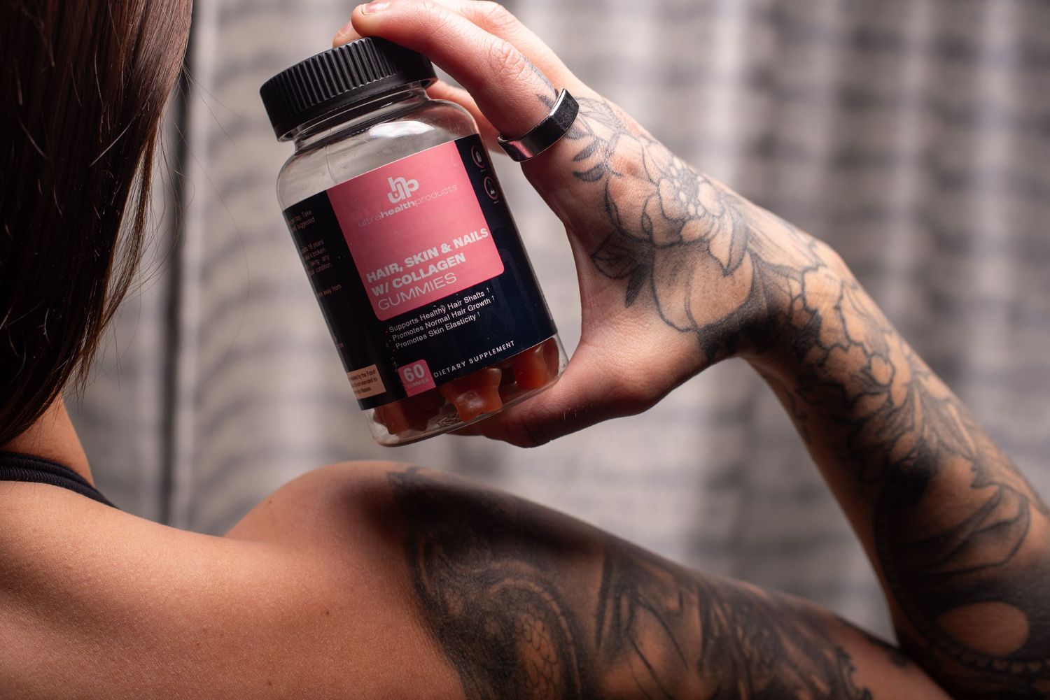Tattooed arm holding up a red and black supplement bottle against a blurred background.