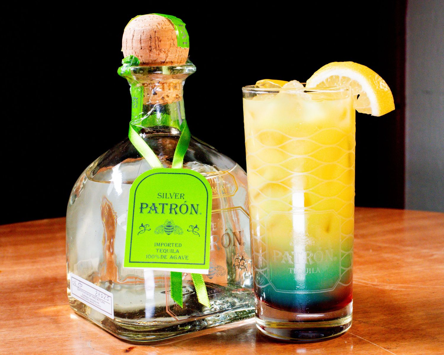 Patron silver tequila bottle next to a layered tropical cocktail with orange garnish on wooden surface.