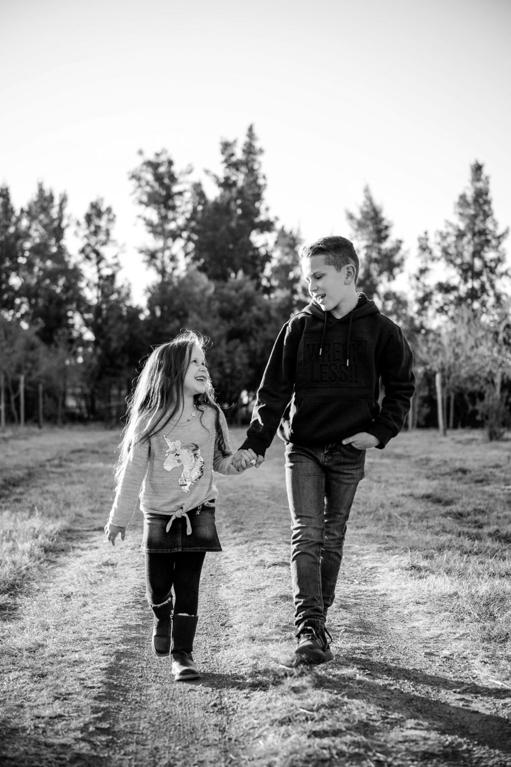 Black and white photo of two siblings walking hand-in-hand down a tree-lined path.
