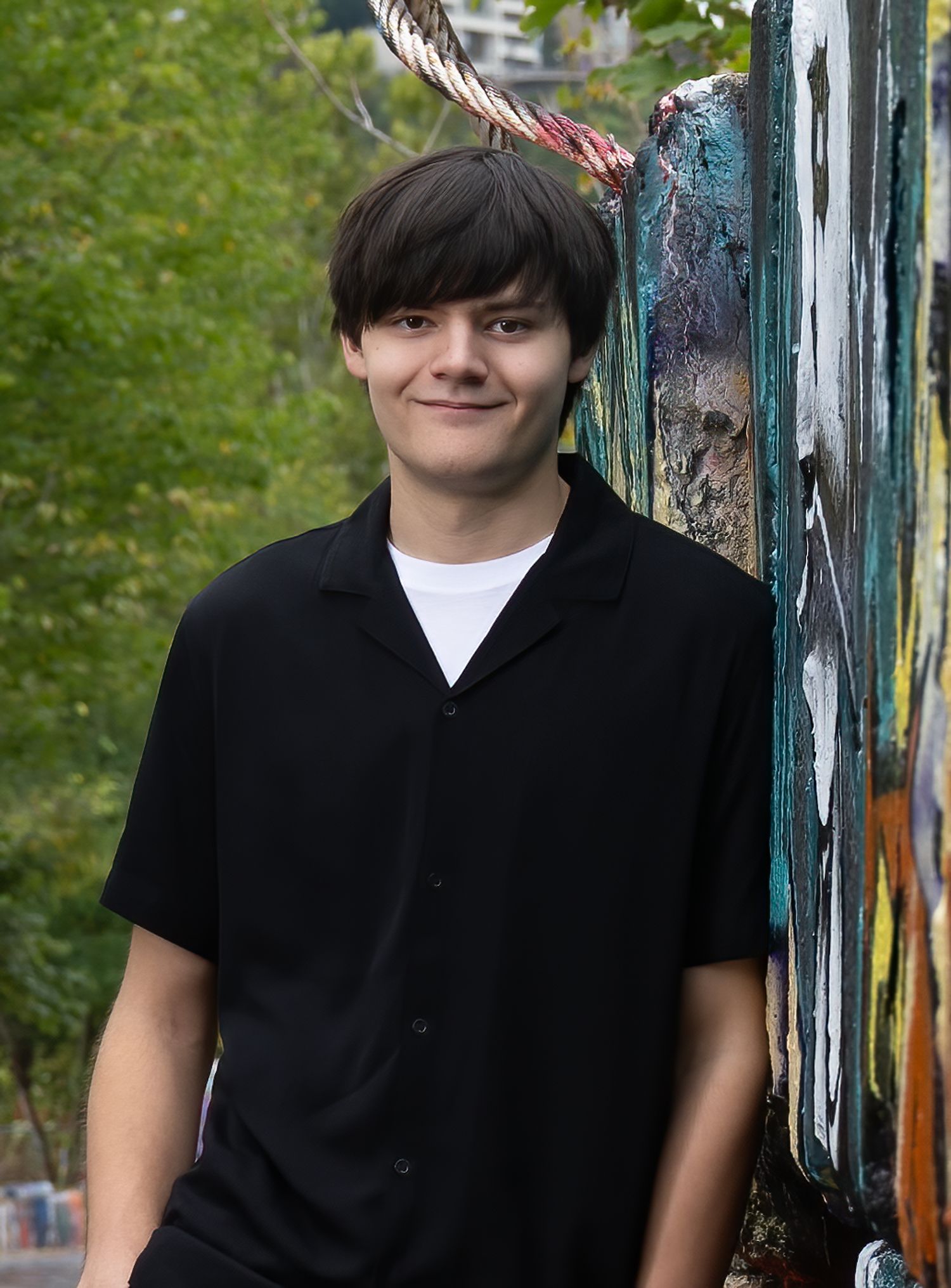 Portrait of a young person in a black button-up shirt standing against a graffiti wall outdoors.