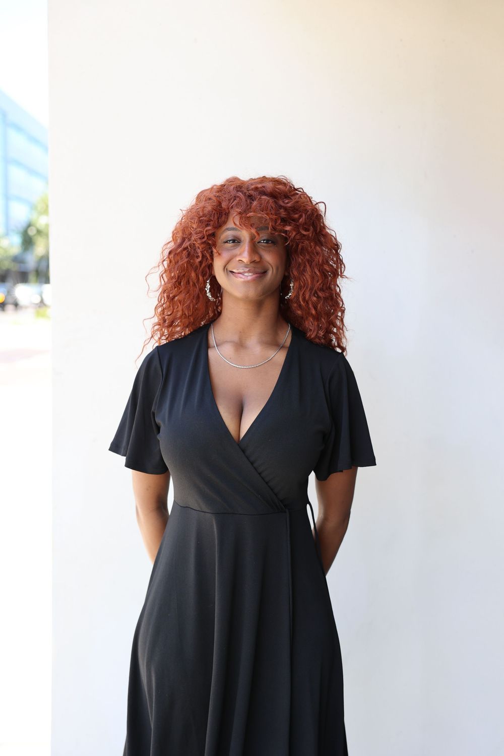 Woman in black dress with curly red hair smiling against white wall.
