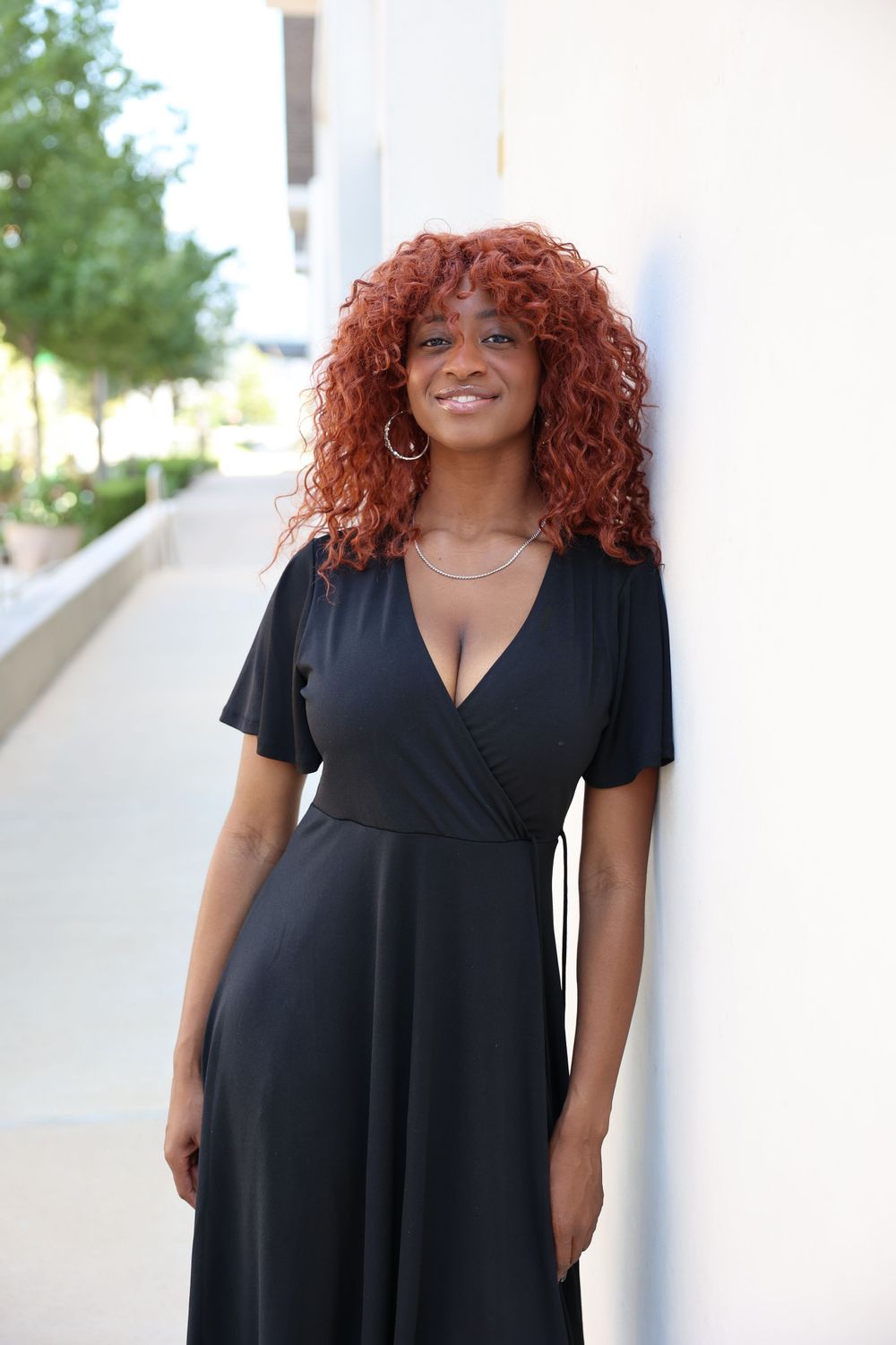 Woman with bright red curly hair wearing black wrap dress poses outdoors.