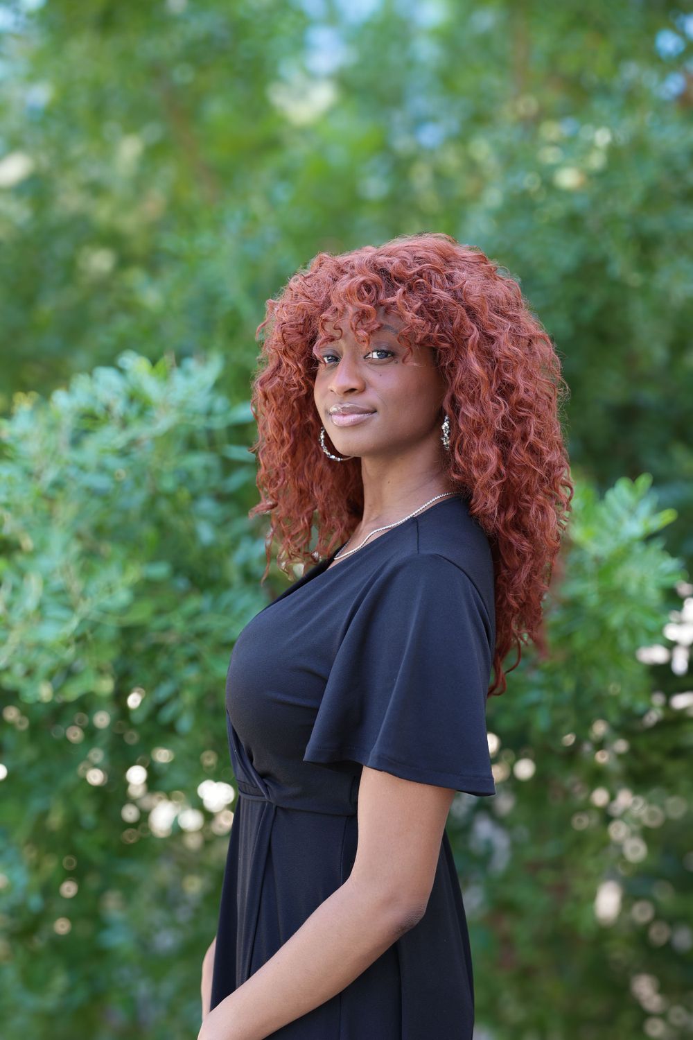 Woman with vibrant red curly hair poses in black dress against blurred green foliage background.