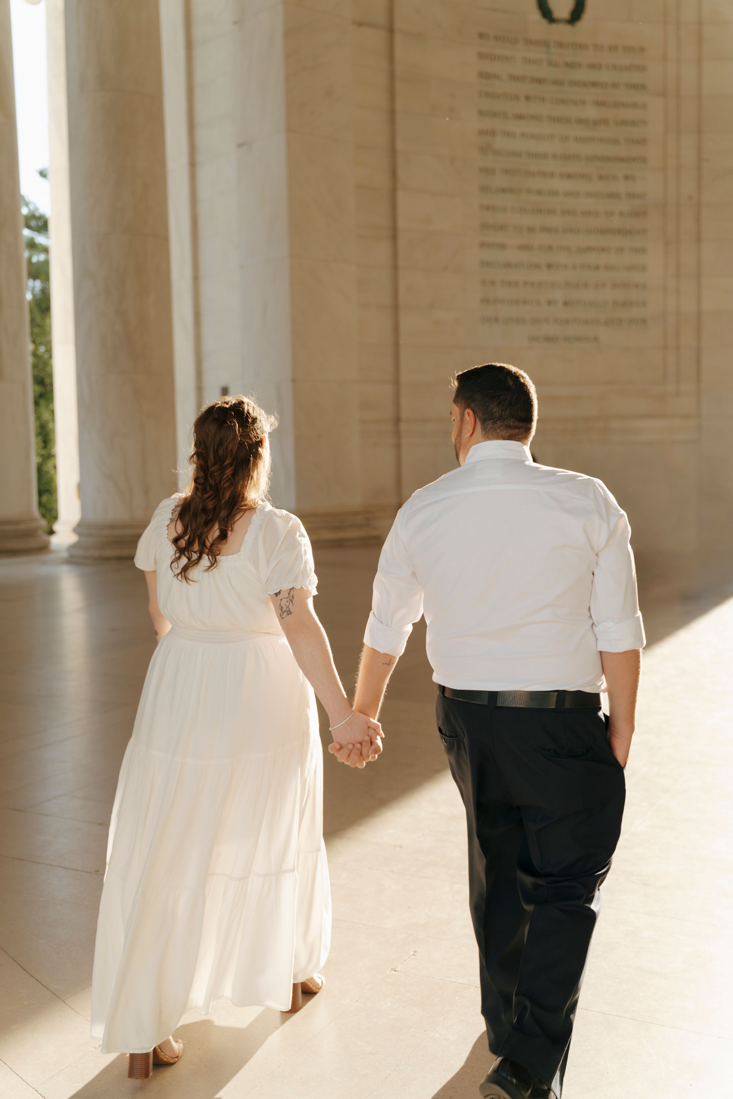 Couple in white attire holds hands walking through Jefferson Memorial columns, viewed from behind.