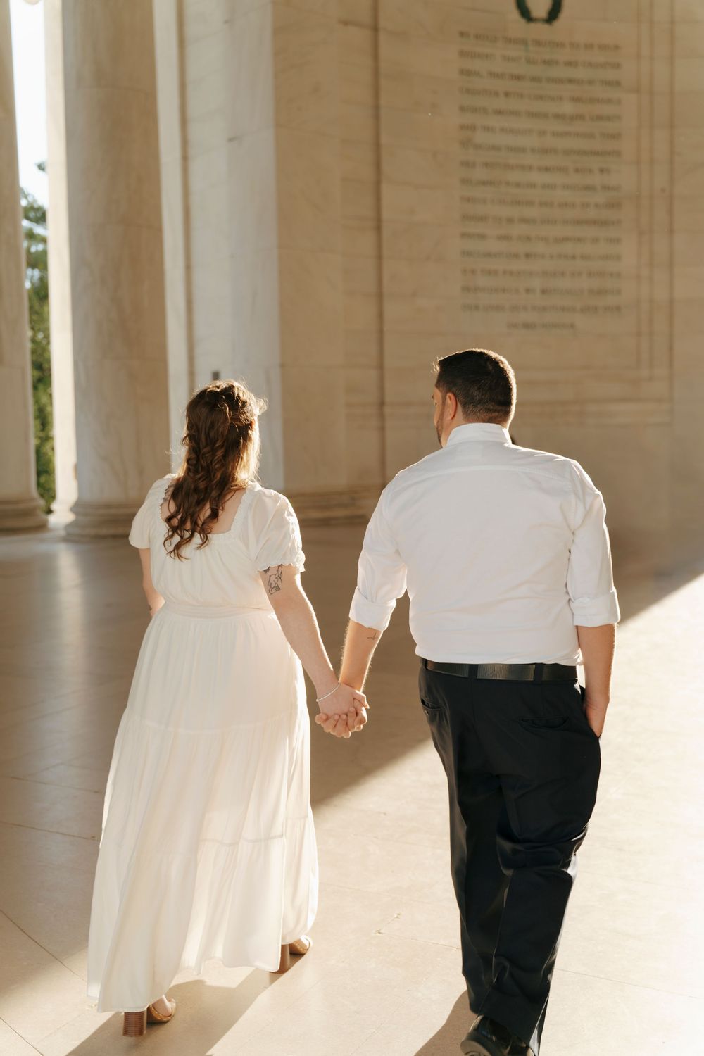 Couple in white attire holds hands walking through Jefferson Memorial columns, viewed from behind.