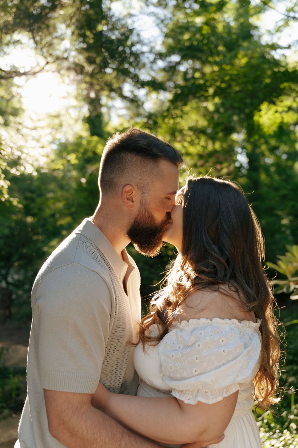 A bearded man and brunette woman share a romantic kiss outdoors, backlit by golden sunlight filtering through green trees.