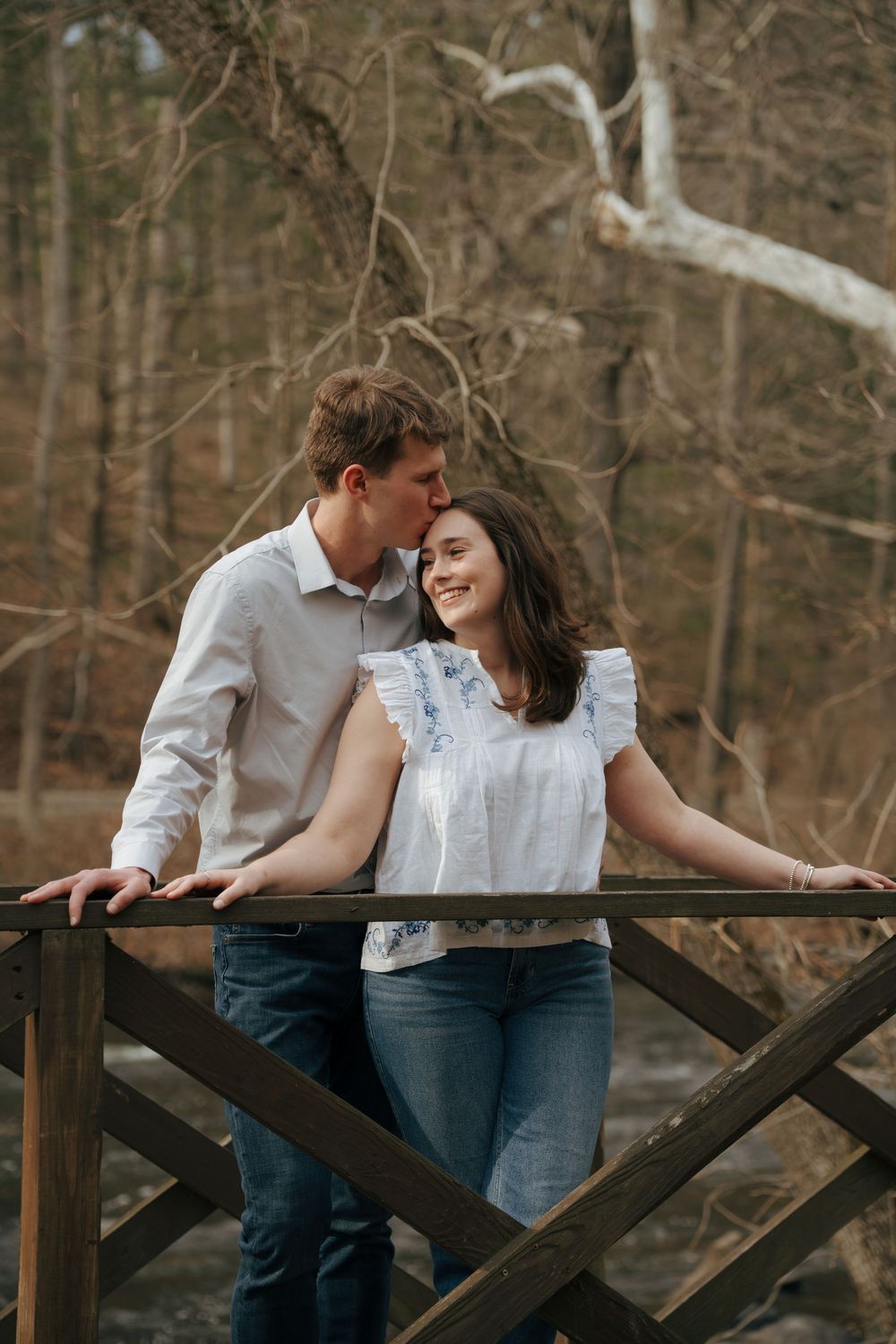 Couple smiling together on a wooden bridge in a forest, woman in white top and man in white shirt and jeans.