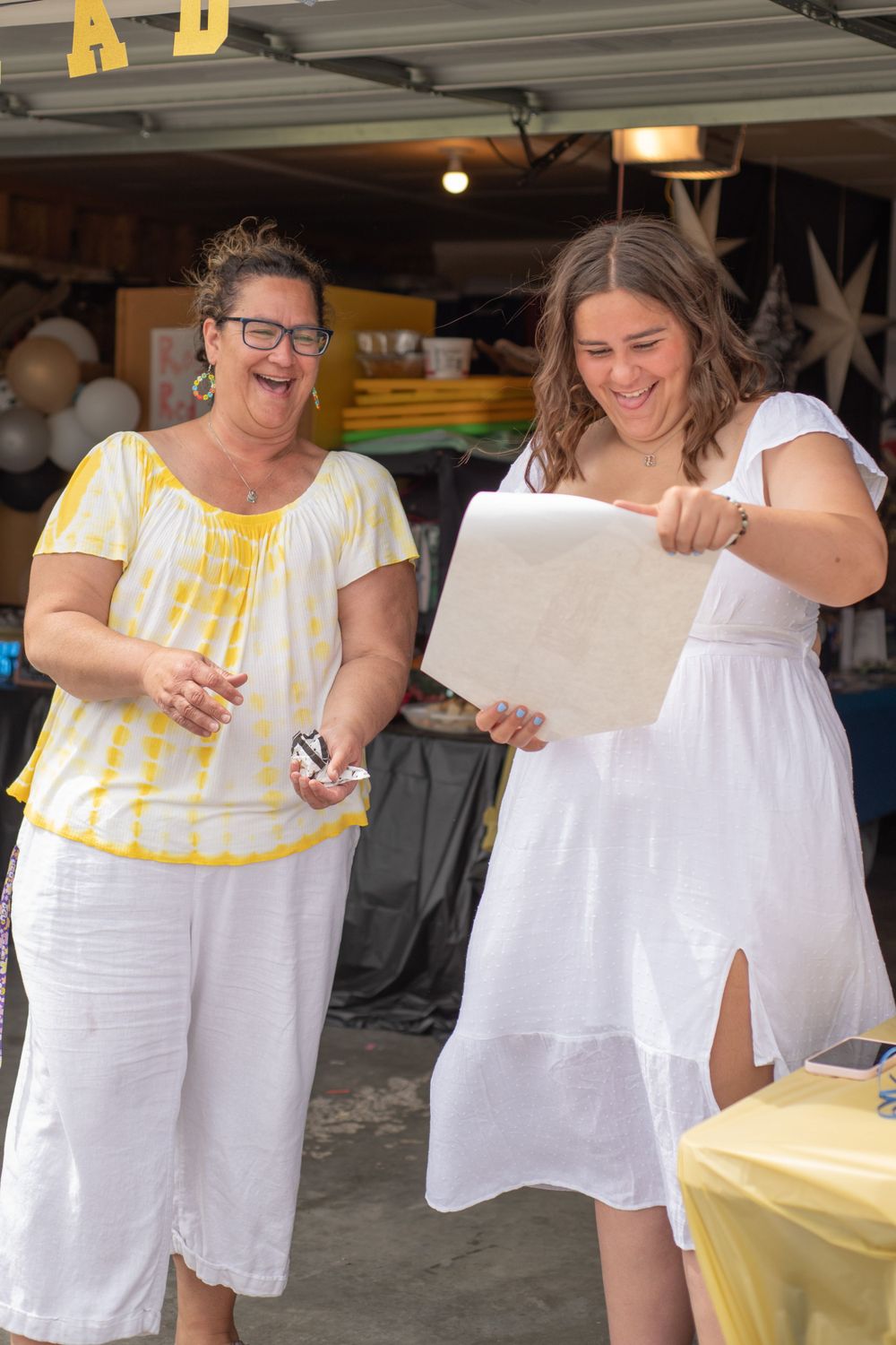 Two women sharing a joyful moment at an outdoor party, one in white dress holding paper.