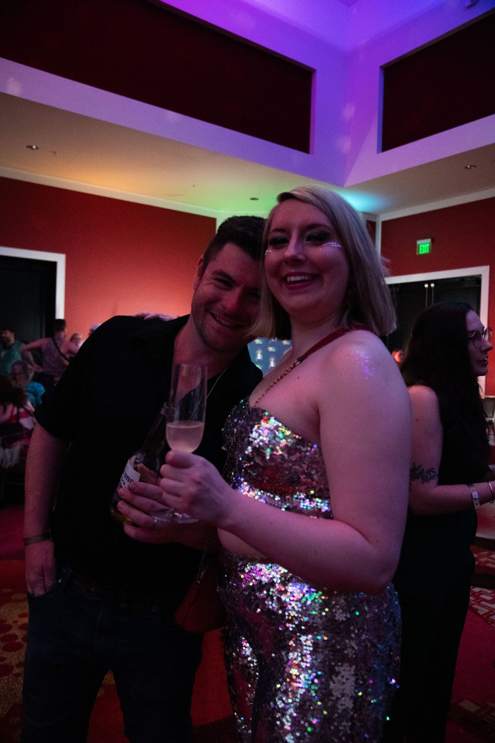 Couple smiling at a party, woman in sequin dress holding champagne glass under colorful lights.