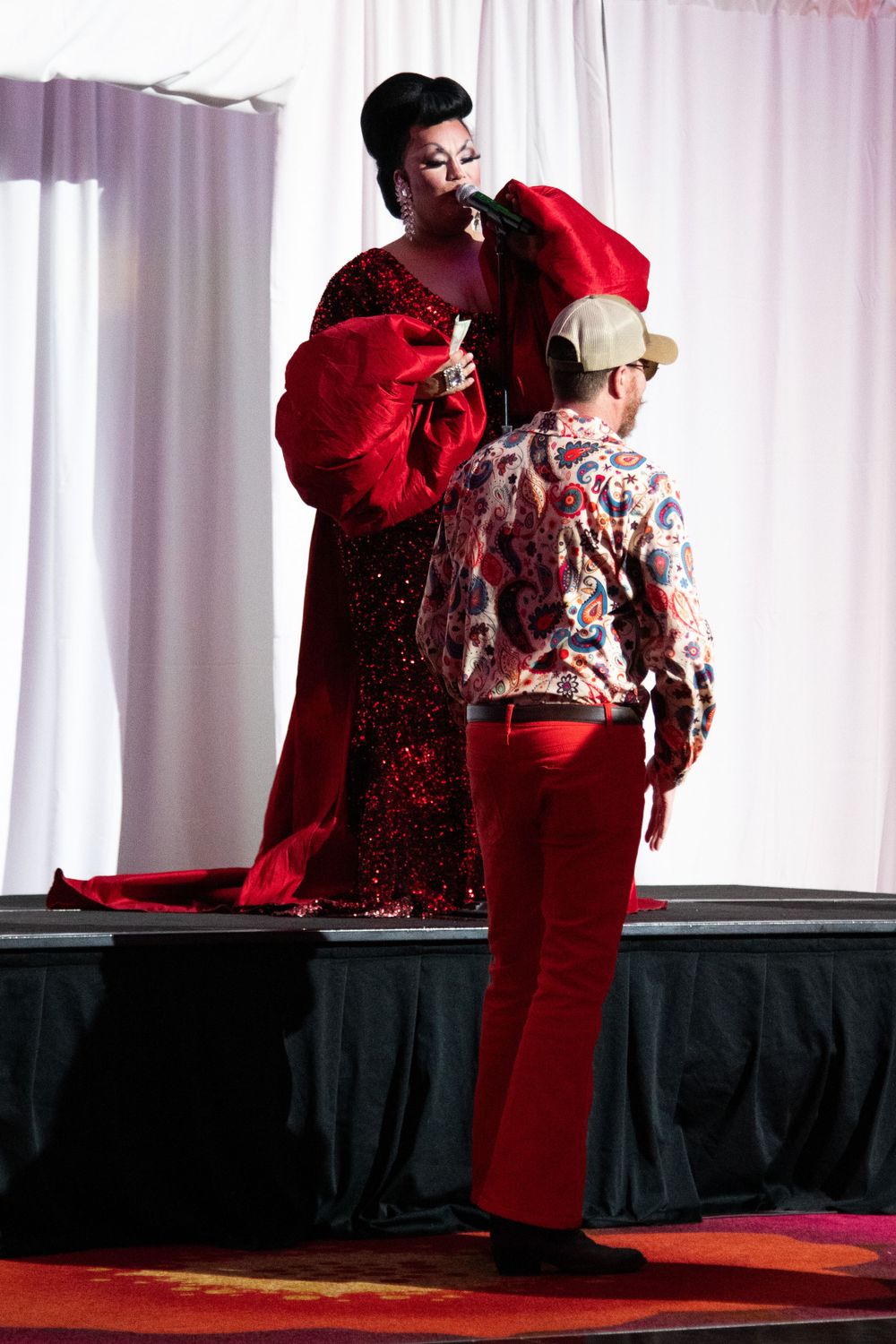 Drag queen in red gown engaging with audience member in red pants on stage.