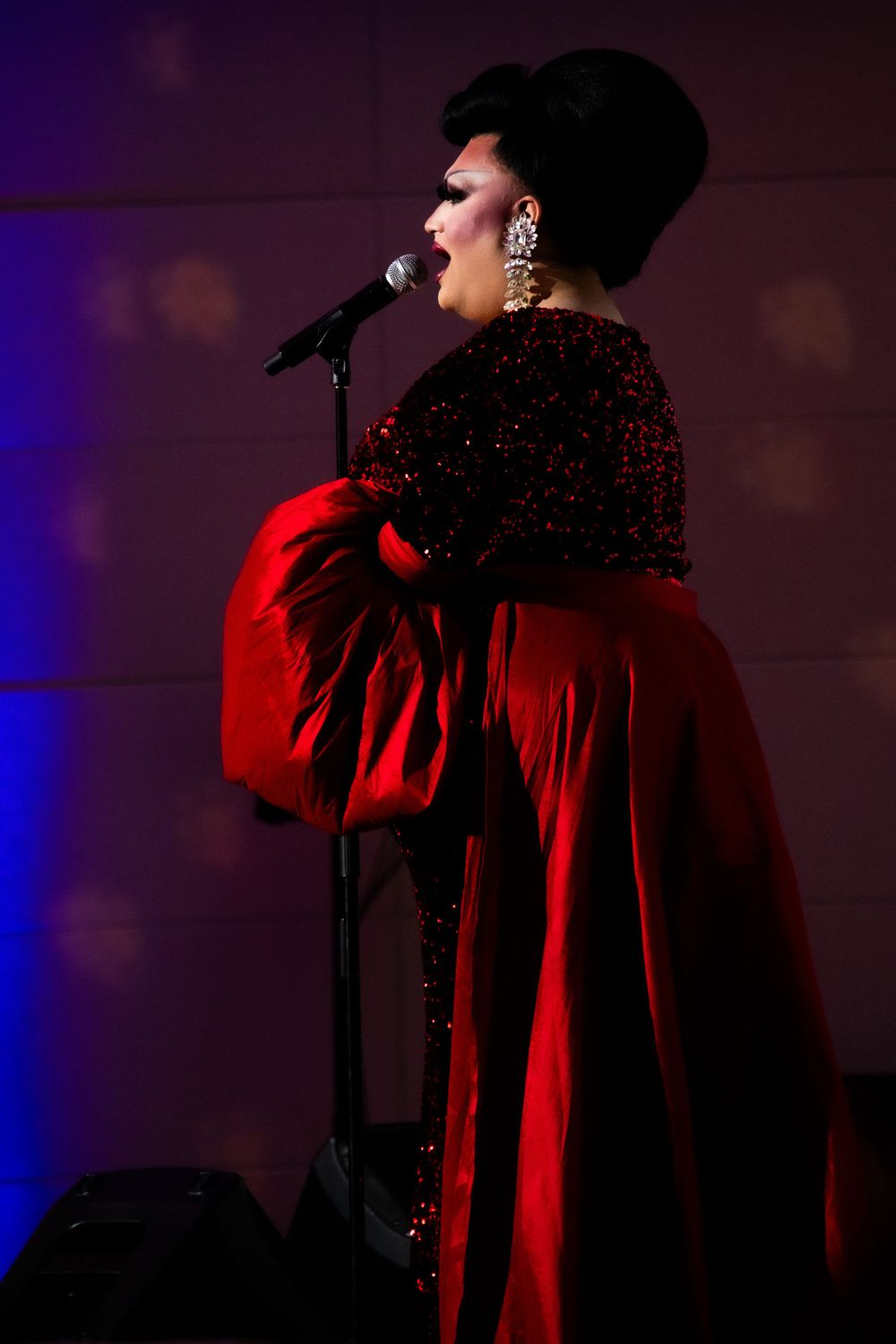 Drag performer in red sequin gown singing at microphone under purple stage lighting.