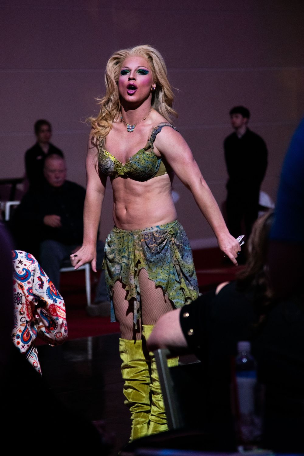 Blonde drag queen in green sequined bra and yellow boots performs on stage before a seated audience.