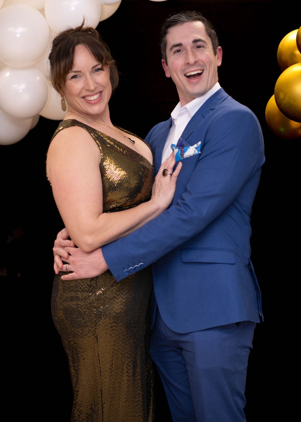 Smiling couple posing together at a formal event, woman in gold dress and man in blue suit with balloons in background.