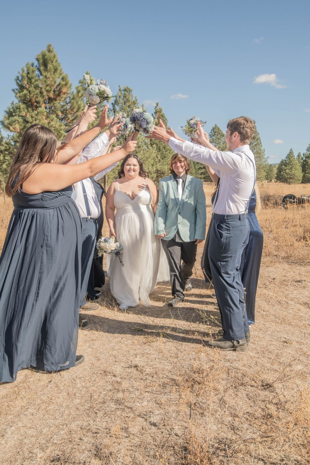 Newlyweds walking through tunnel of raised bouquets created by smiling wedding party in golden field.