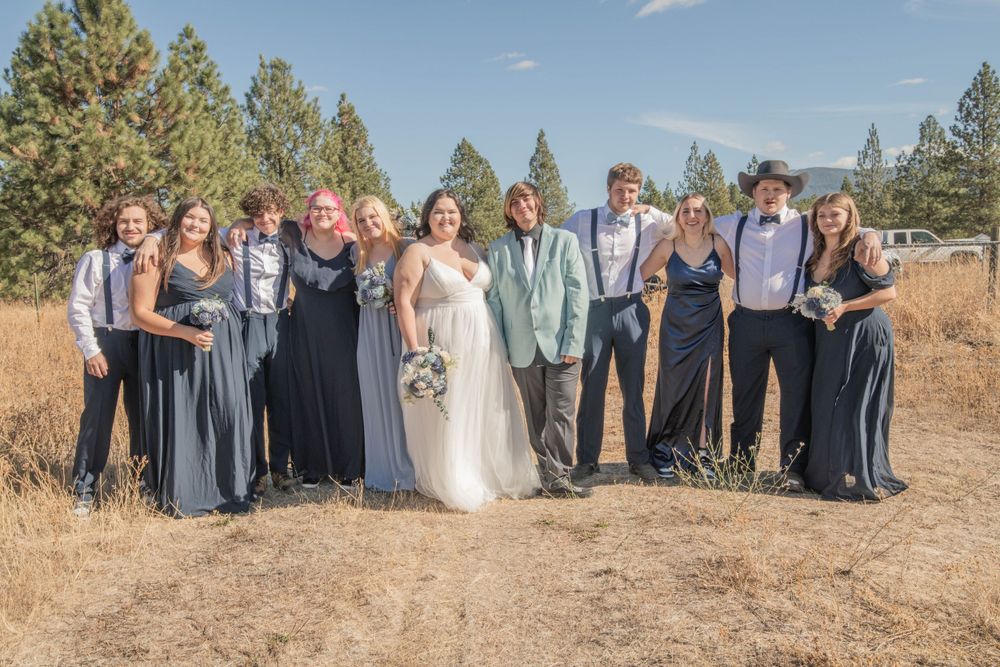 Bride and groom with full wedding party in blue gowns and suspenders posing in outdoor rustic setting.