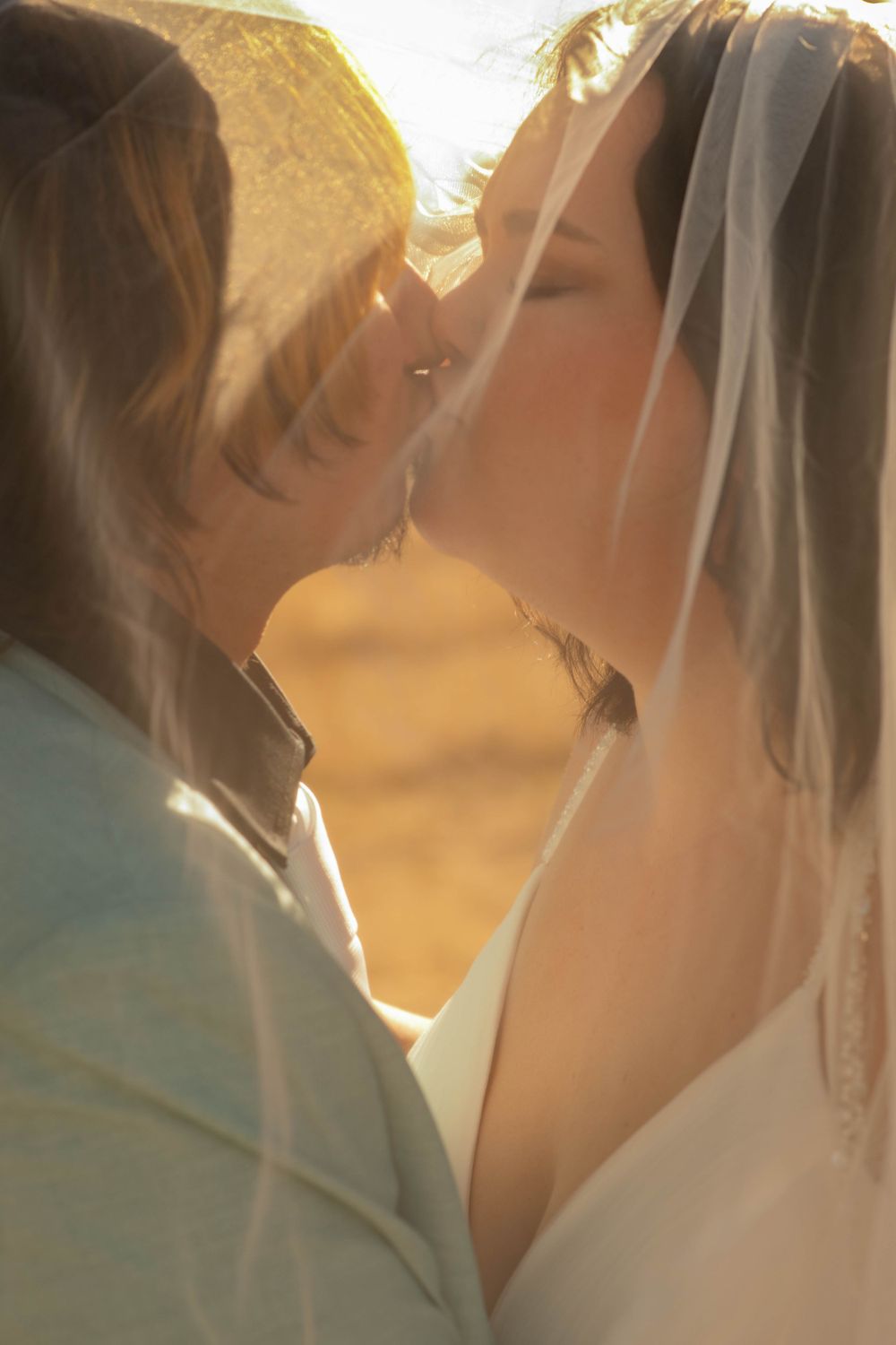 Romantic bride and groom kissing under sheer wedding veil in warm golden sunset light.