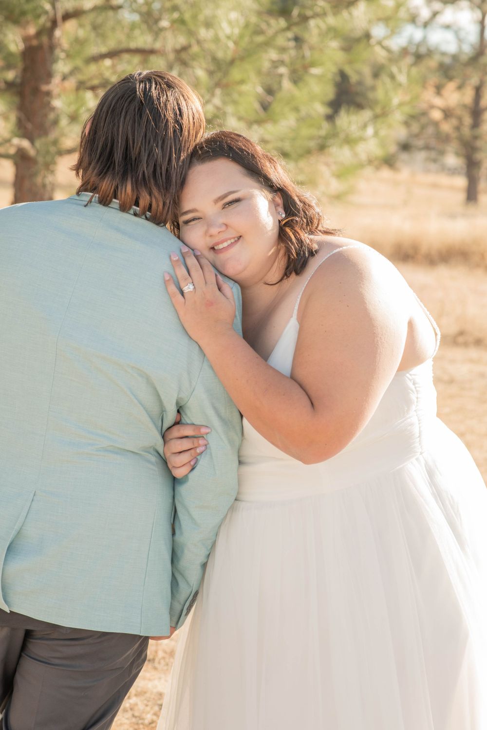 Smiling bride in white wedding gown embracing groom from behind near pine trees on wedding day.