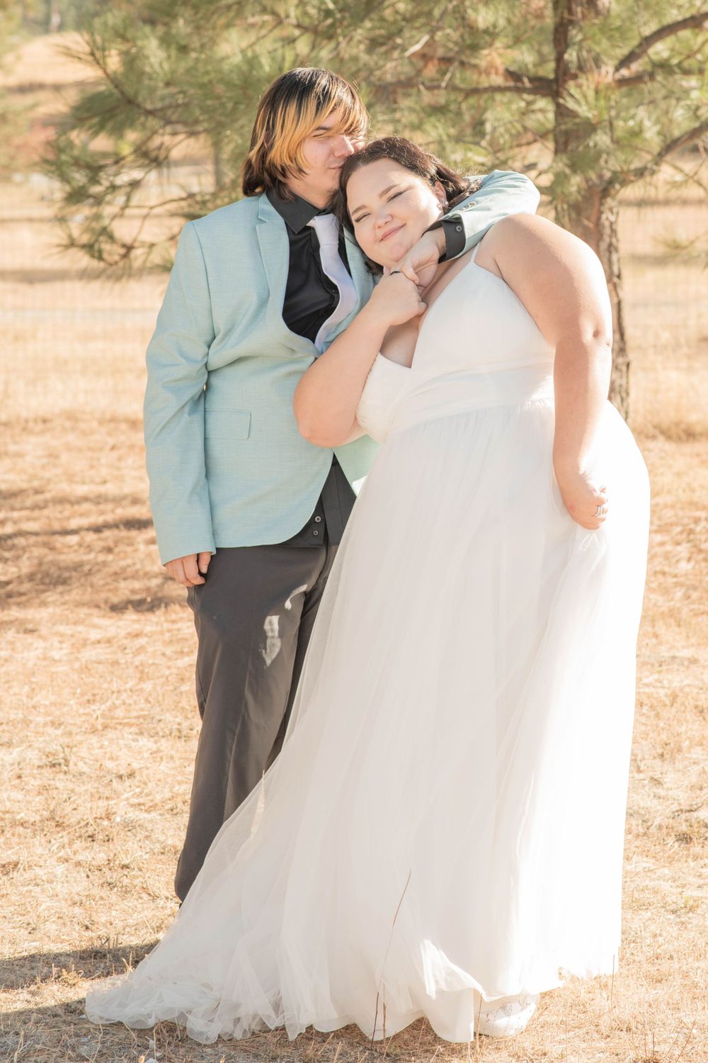 Happy bride and groom posing together outdoors, groom in mint blazer hugging bride in white dress.
