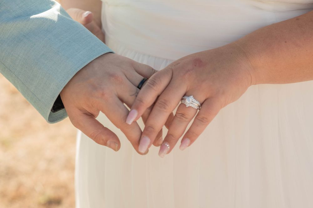 Close-up of newlyweds holding hands displaying sparkling wedding rings against white bridal gown.