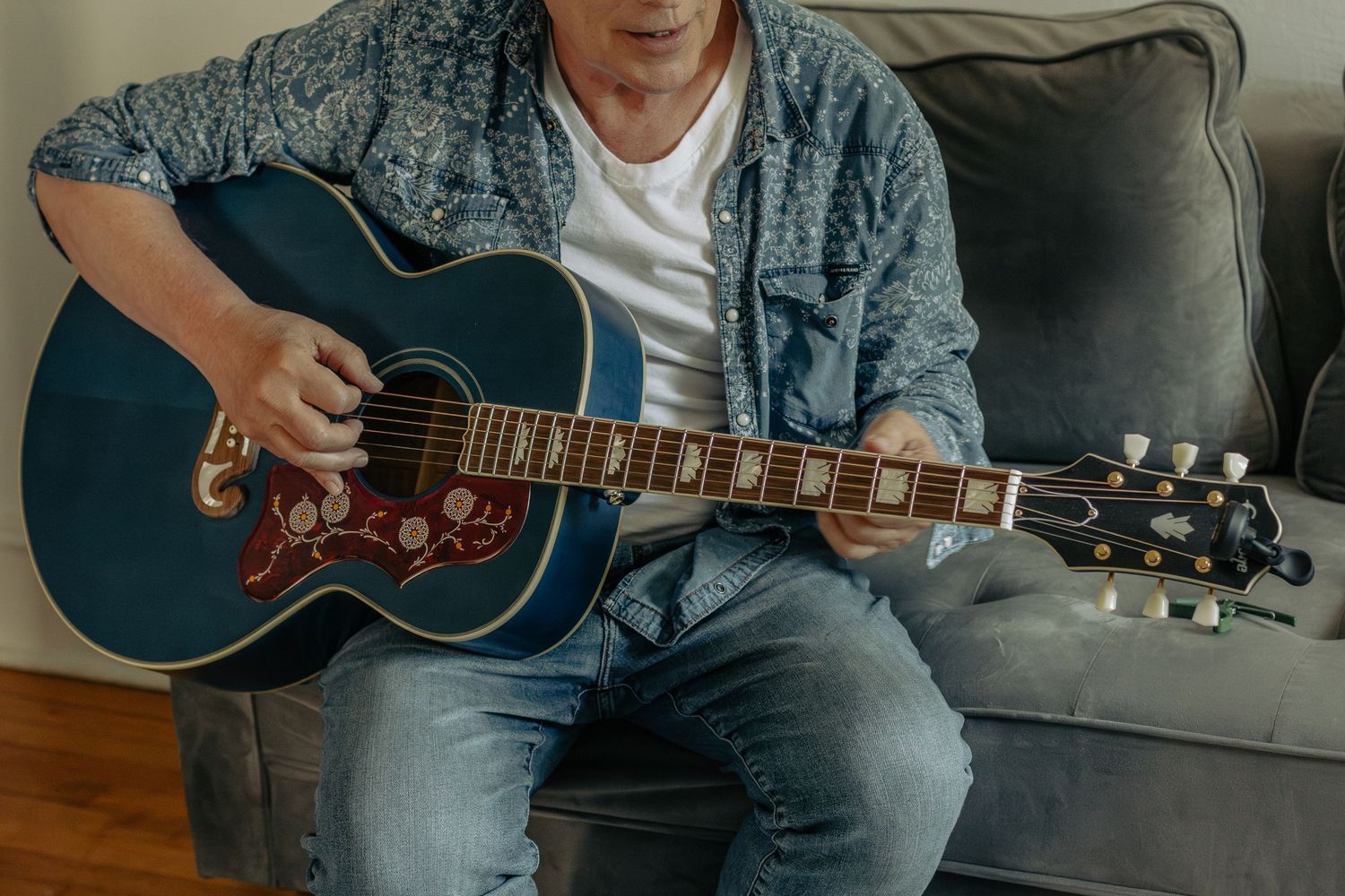 Person playing blue acoustic guitar while sitting on grey couch in casual denim outfit.