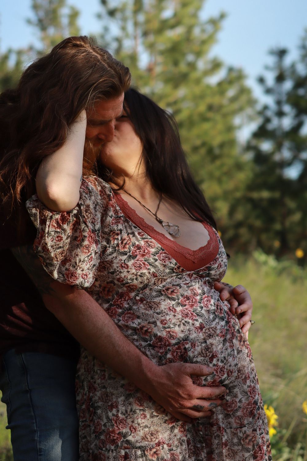 Romantic outdoor maternity photo showing a couple embracing, with woman wearing floral dress against natural backdrop.