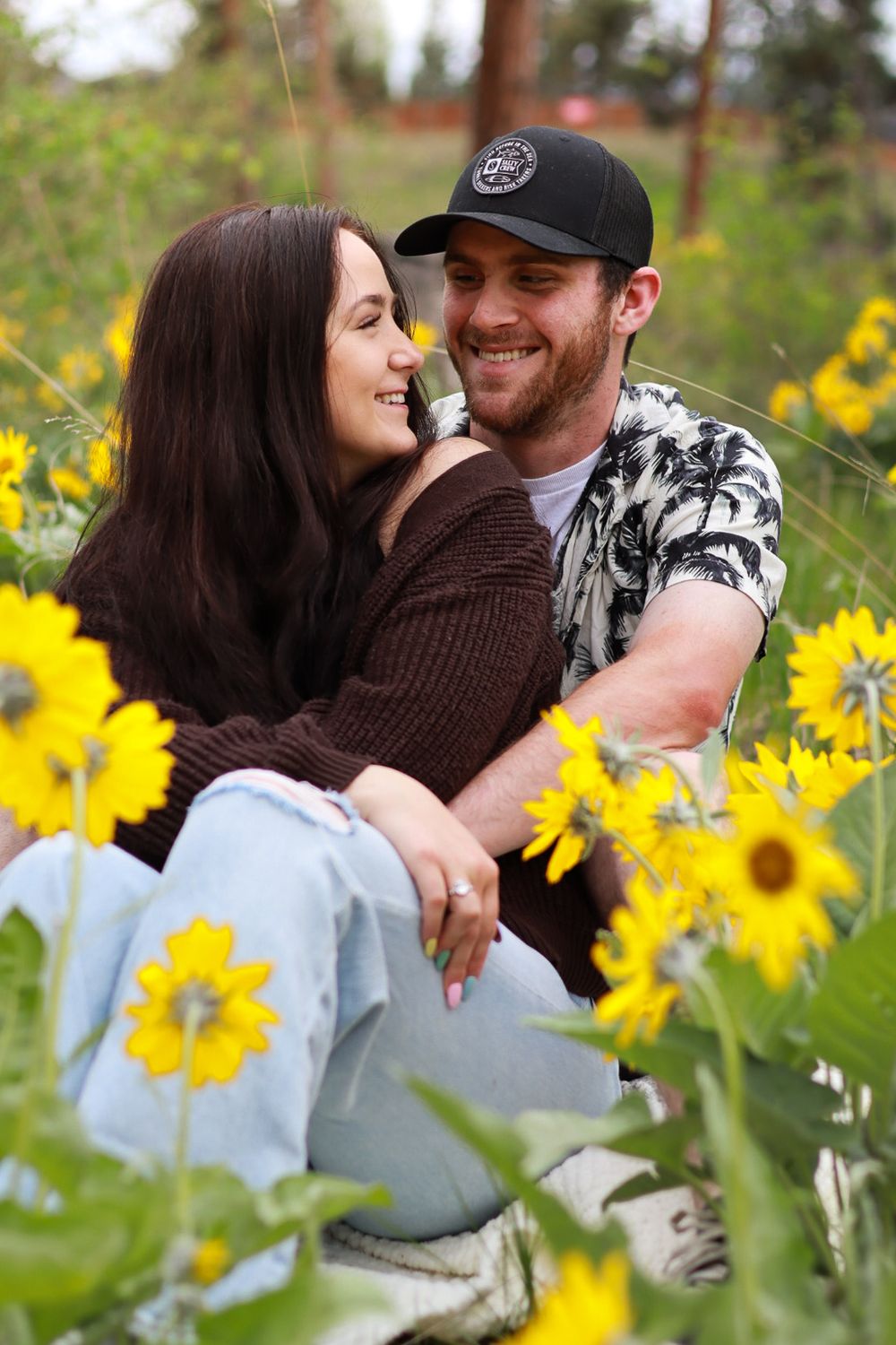 A couple shares an intimate moment while sitting among blooming sunflowers in a garden setting.