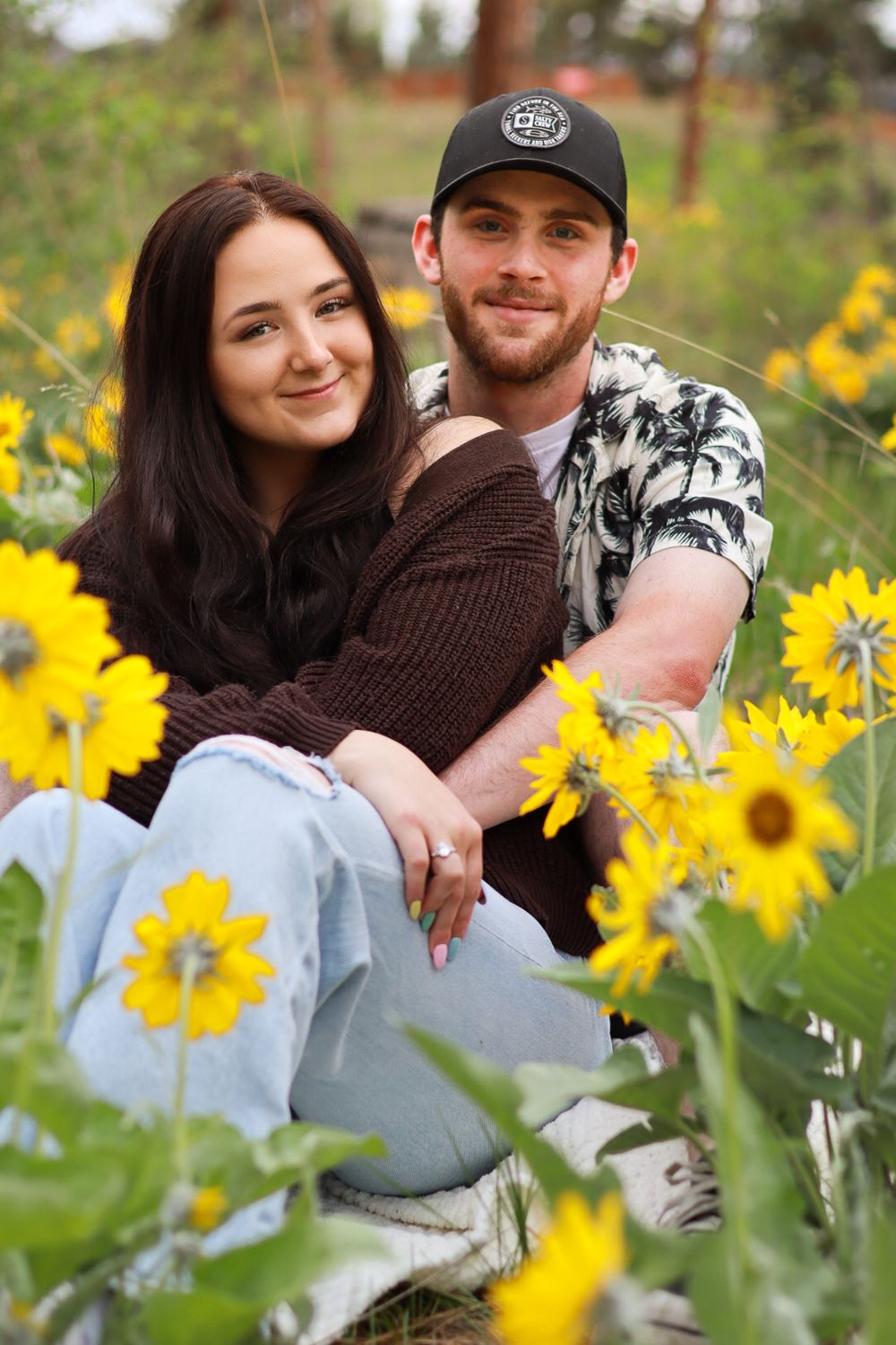 A couple sits embraced among bright yellow sunflowers in a garden setting on a sunny day.