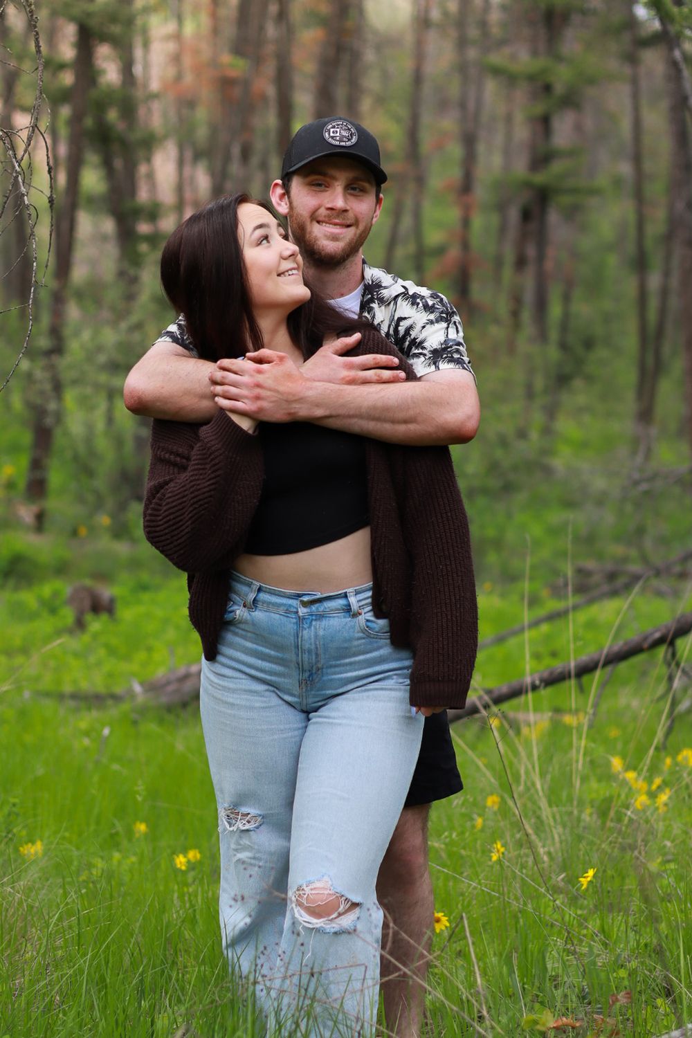 A couple embraces while standing in a forest meadow with spring wildflowers and green grass.