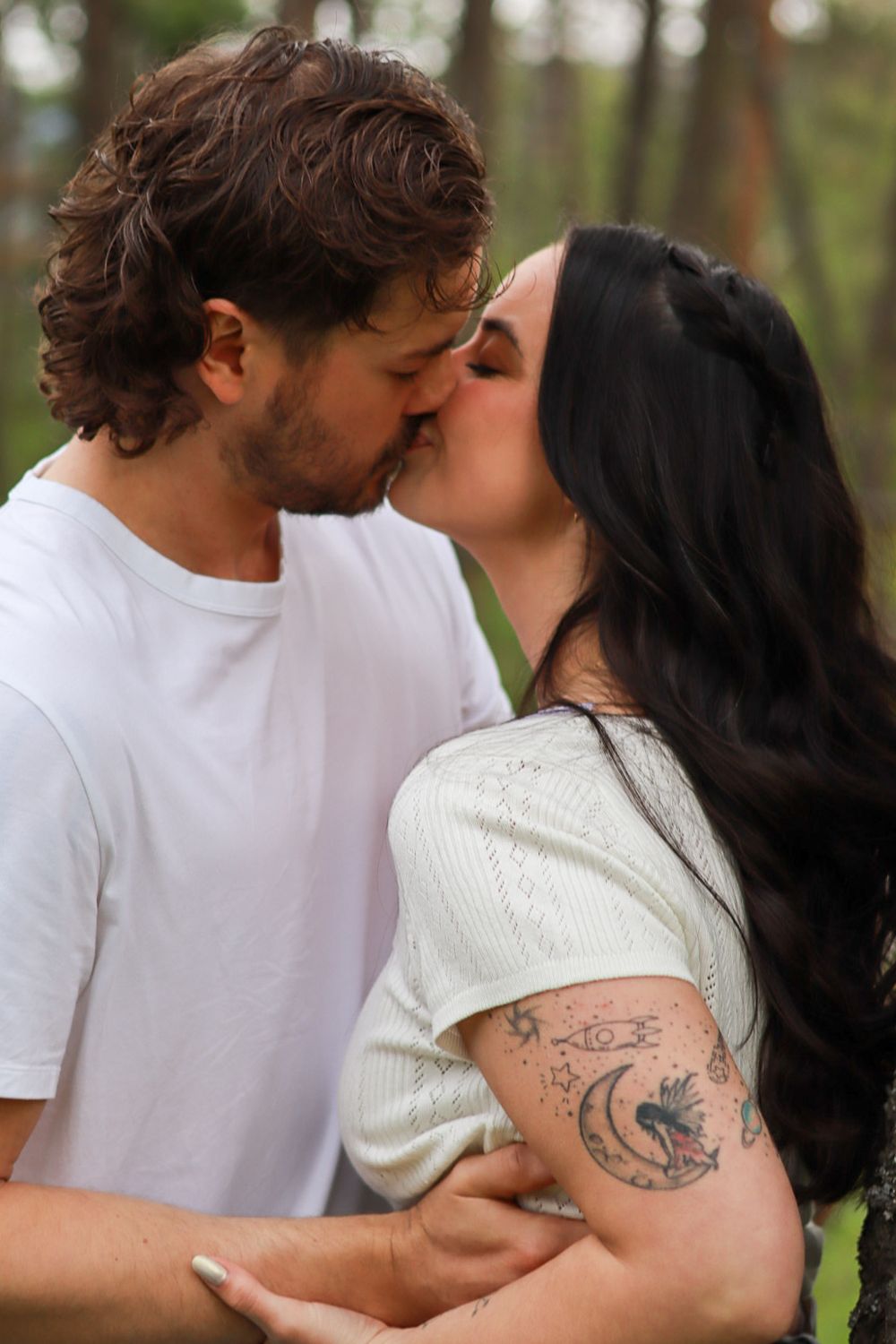 A couple wearing white embraces in a tender kiss outdoors with a visible arm tattoo in the foreground.