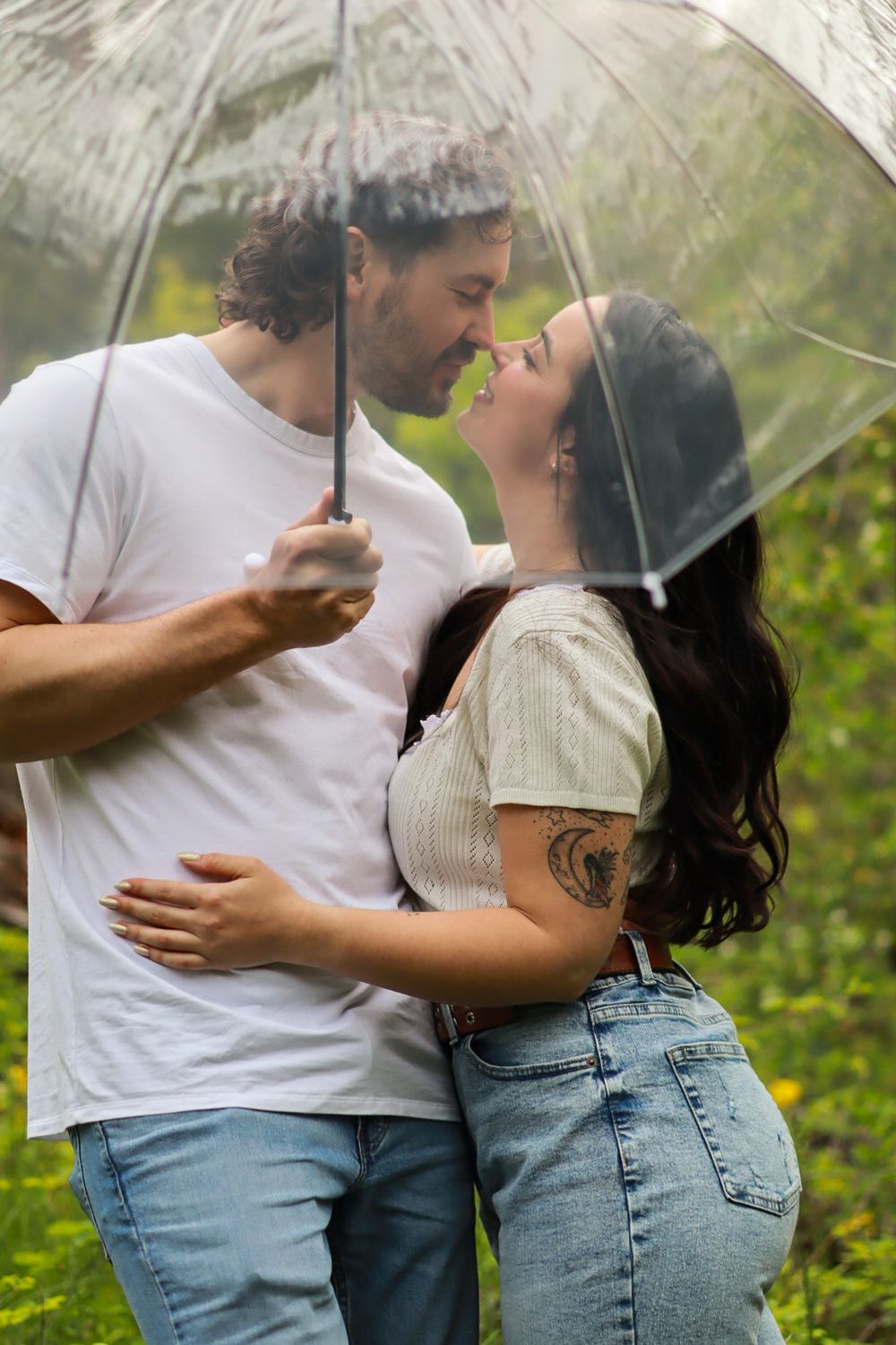 Romantic couple sharing an intimate moment under a clear umbrella in a lush green setting.