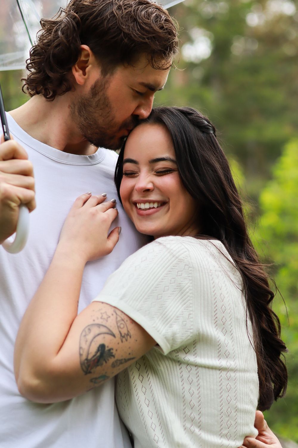 A couple embraces under an umbrella outdoors, sharing a tender romantic moment in casual white clothing.