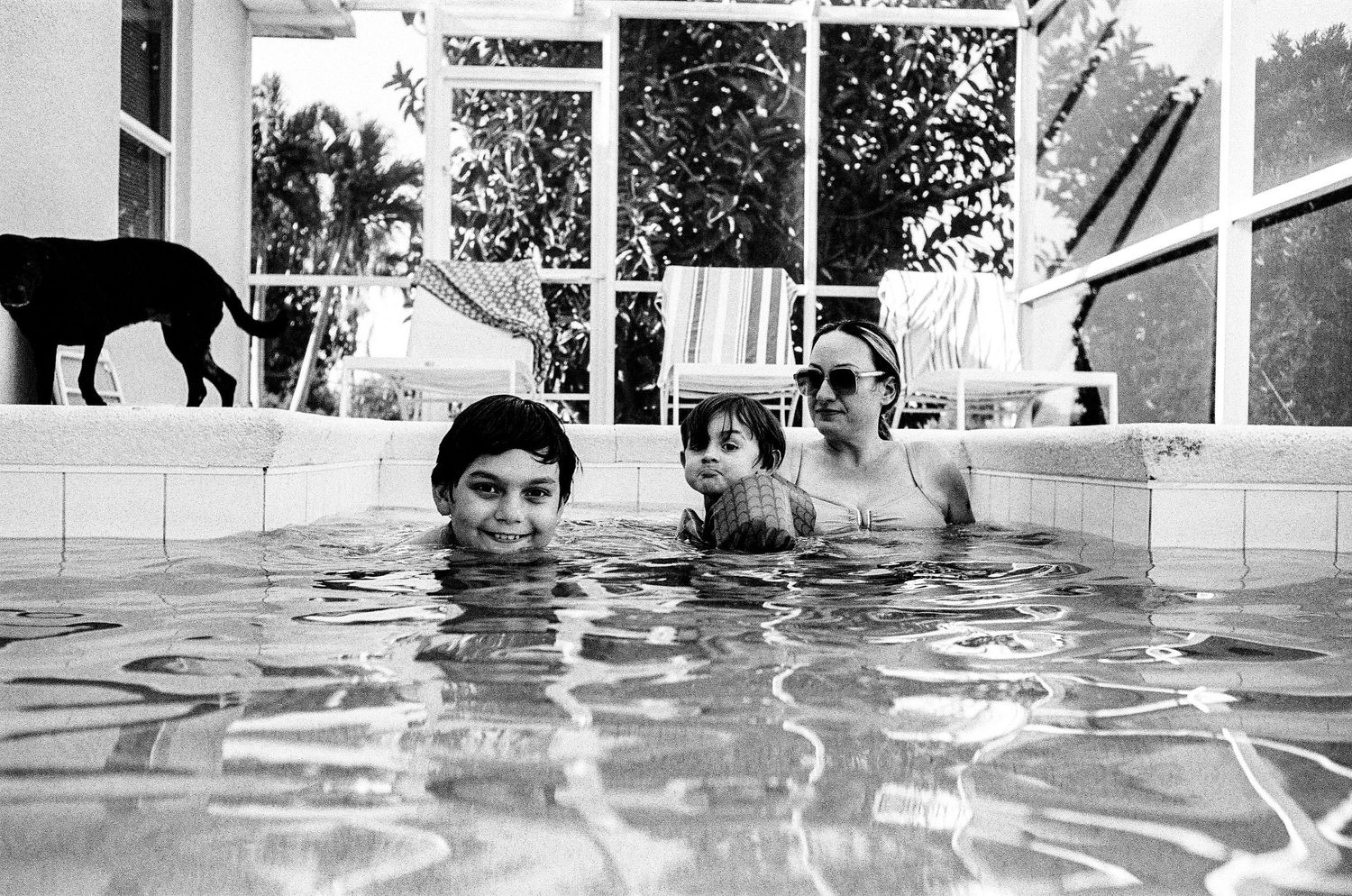 Three people enjoying a swim in a pool while a black cat walks along the edge in black and white photography.