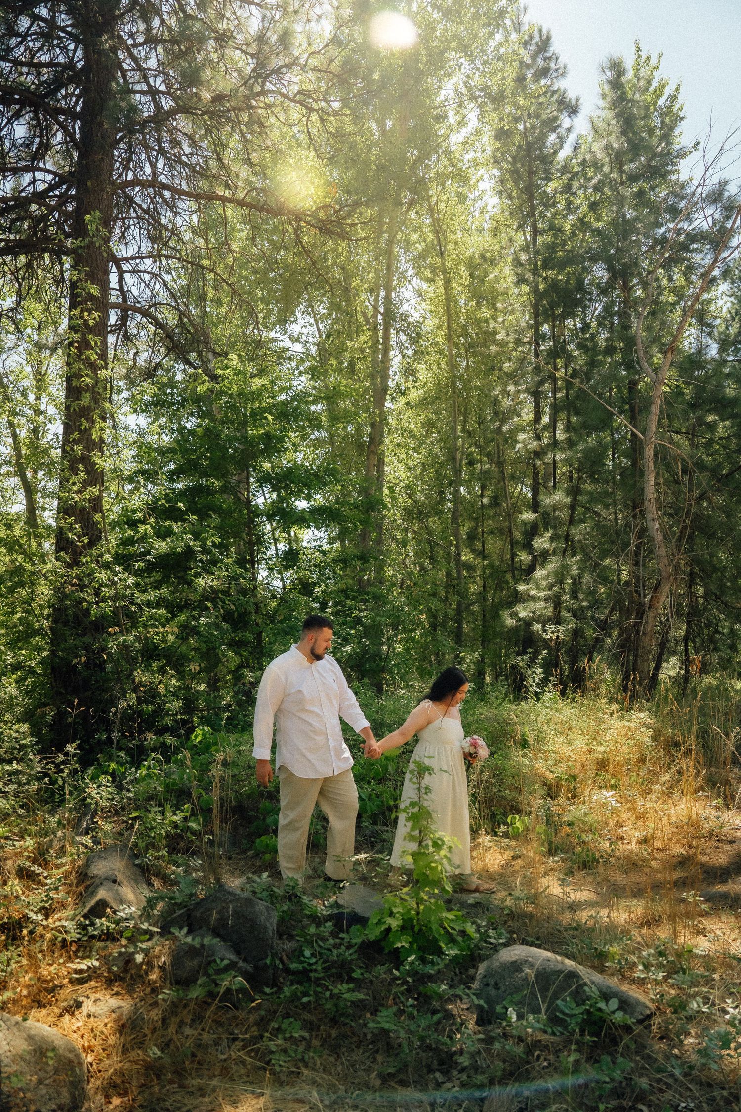 Couple holding hands while walking through a sunlit forest clearing on a bright summer day.
