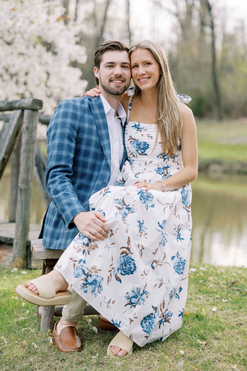 A couple poses outdoors by a pond, with her wearing a floral dress and him in a blue plaid blazer in Warwick, New York.