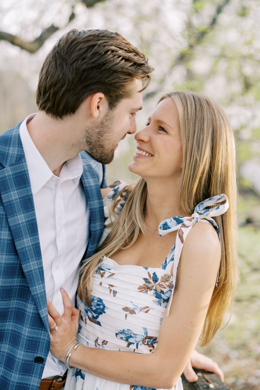 A romantic spring portrait of a couple embracing among cherry blossoms, with her in a floral dress and him in a blue blazer in Warwick, NY.