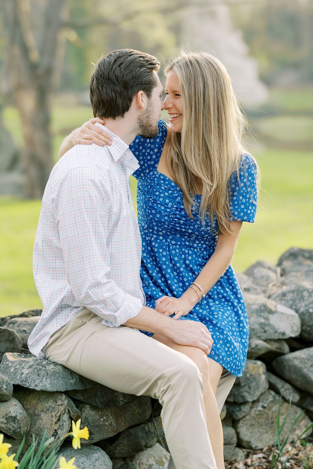 A couple shares an intimate moment sitting on a stone wall outdoors with daffodils blooming nearby in spring in Warwick, New York.