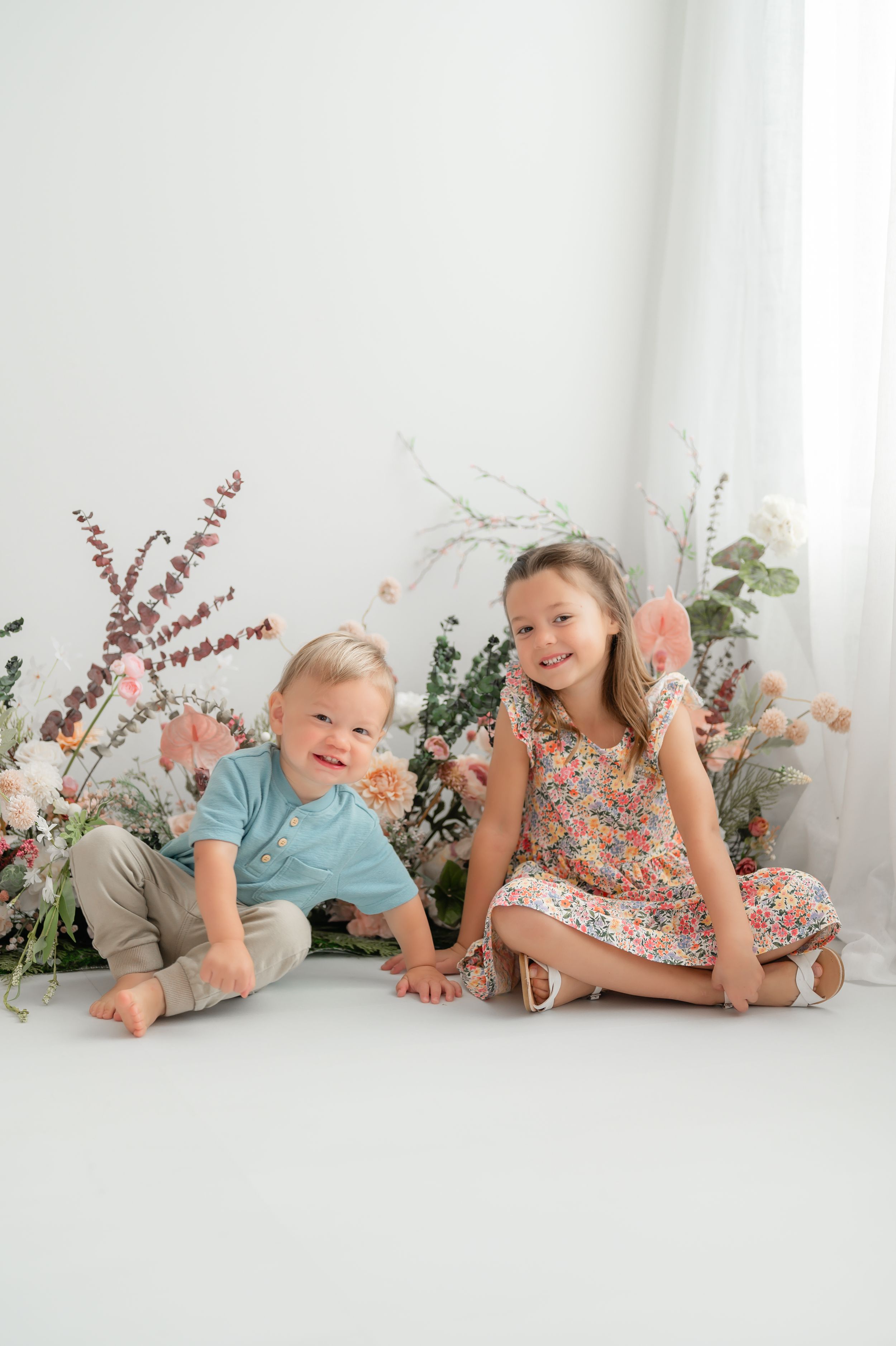 Two young children sit against a white backdrop with delicate floral arrangements featuring greenery and dried flowers.