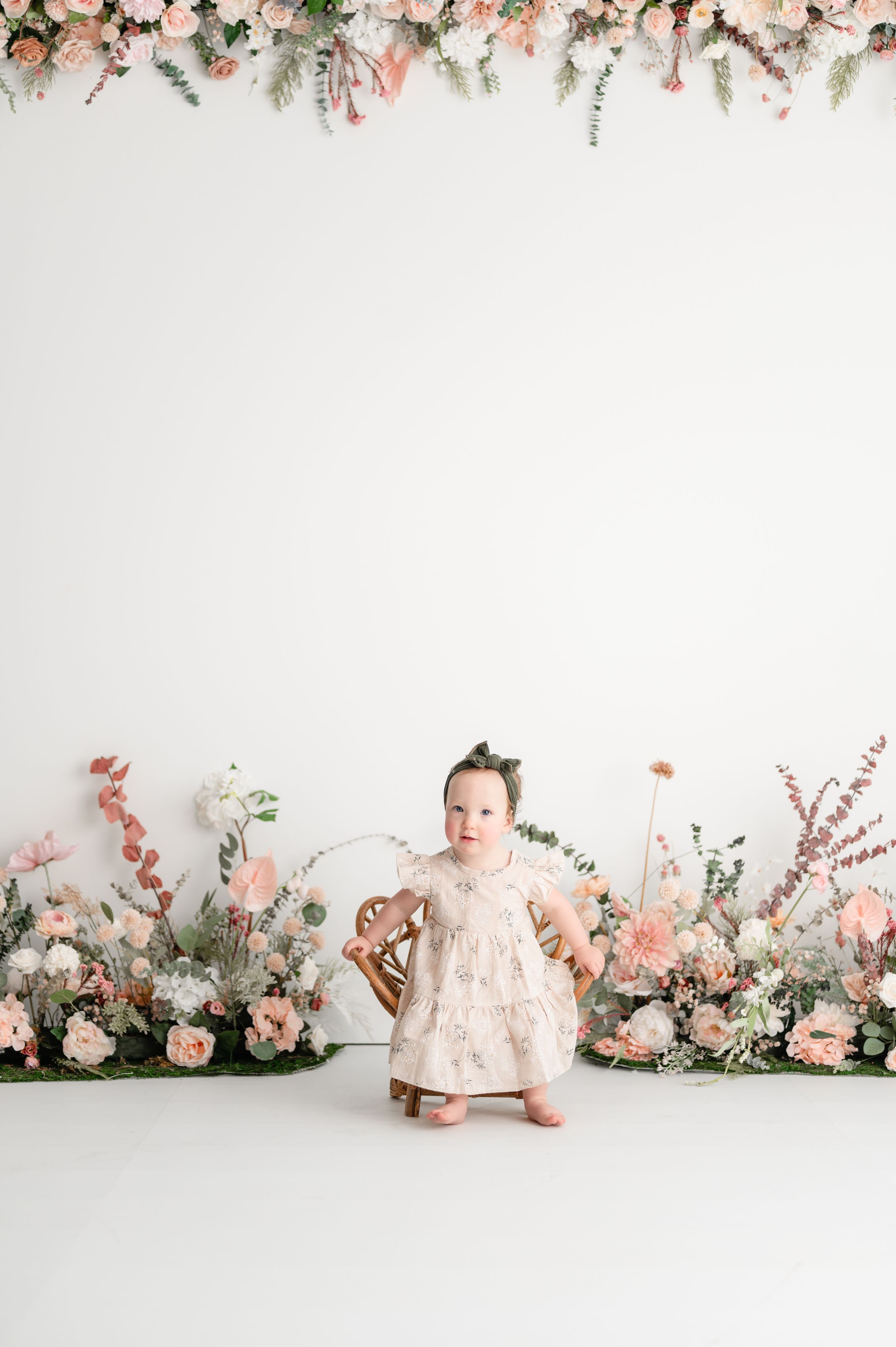 A small child stands among romantic pink floral arrangements against a white backdrop in a photography studio setting.