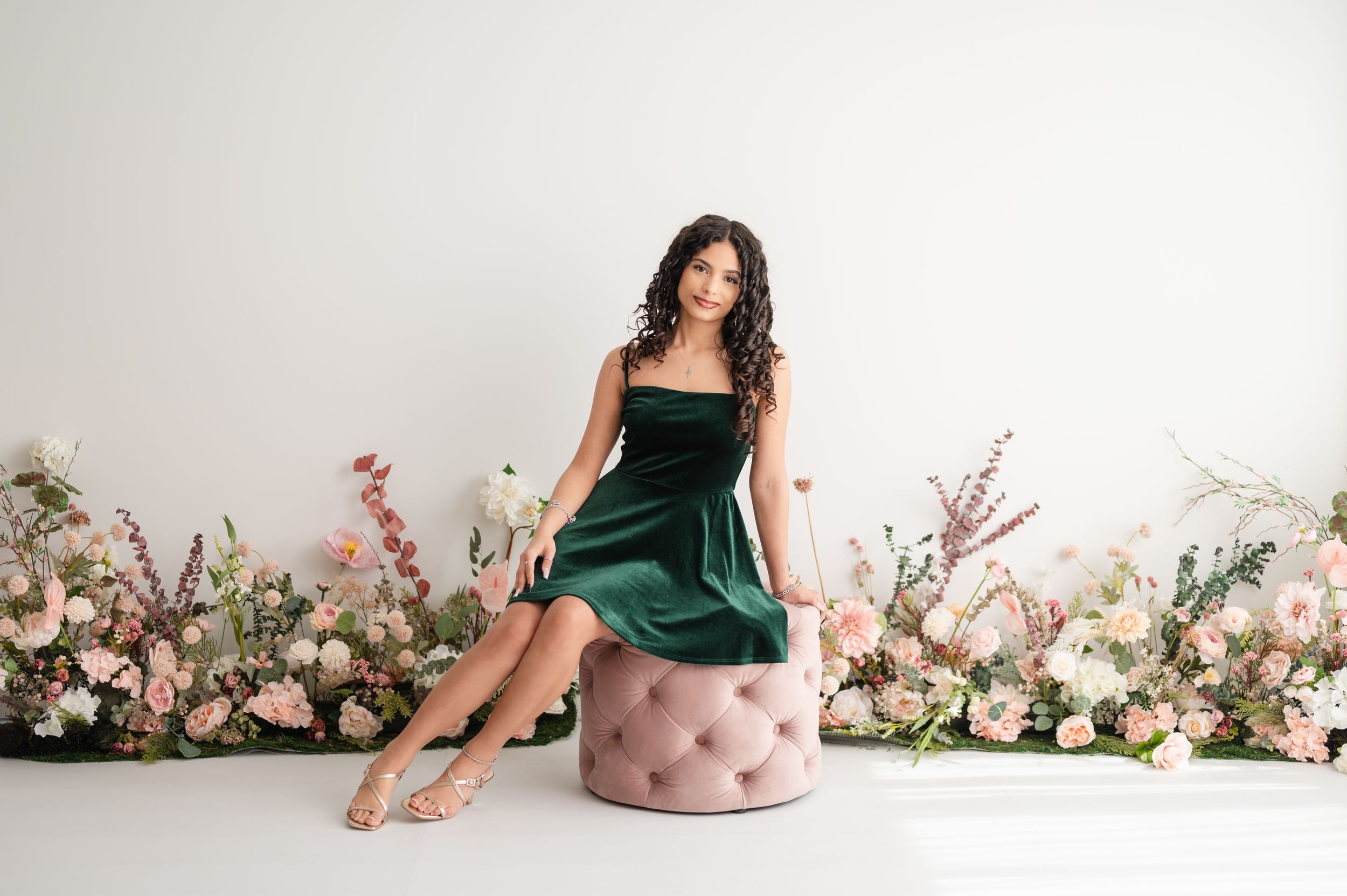 A woman in an emerald green velvet dress poses on a pink ottoman with a backdrop of pink and white floral arrangements.