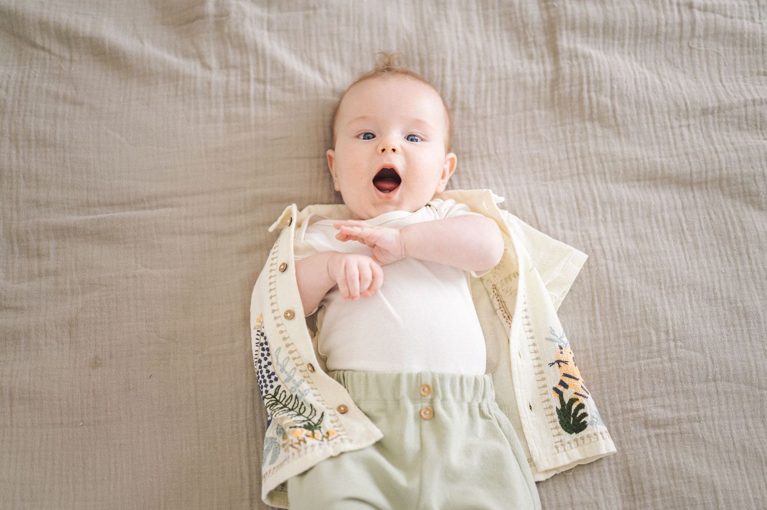 Baby lying on beige bedding wearing a white onesie and pale green pants with an open-mouthed expression.