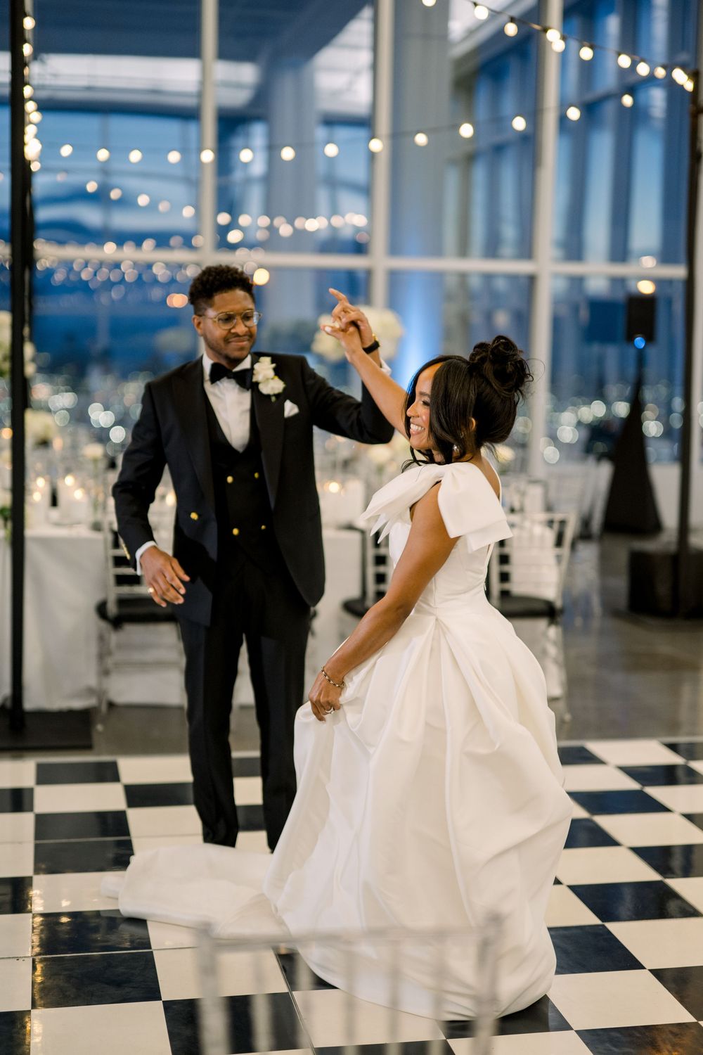 A bride and groom dance together on a black and white checkered floor under twinkling string lights at their wedding reception.