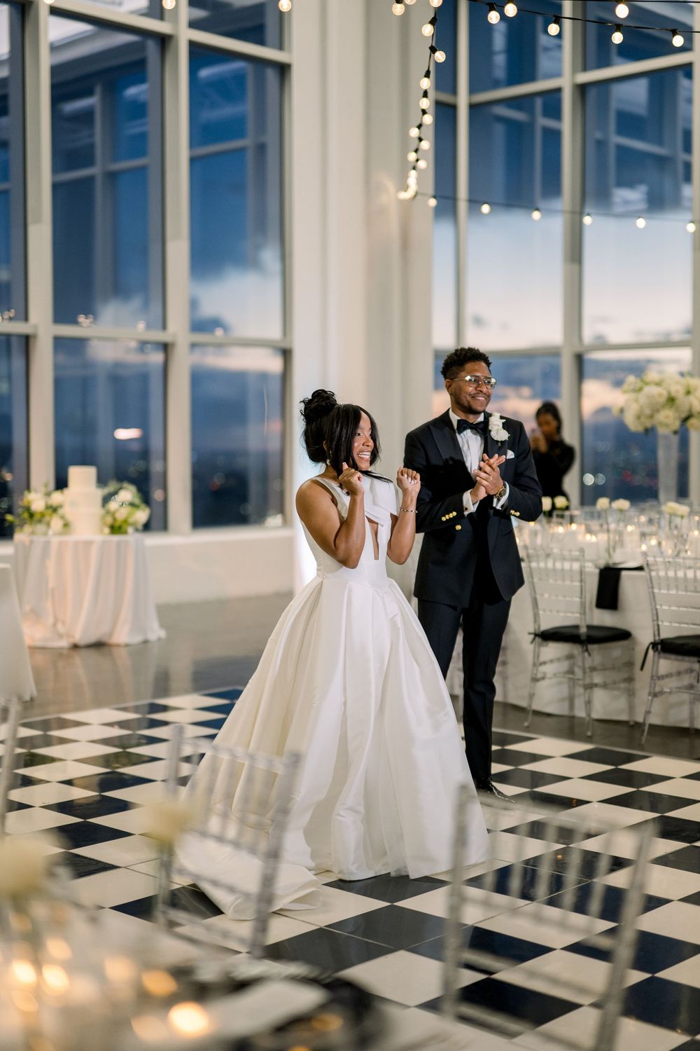 Couple sharing a toast on black and white checkered floor during wedding reception with string lights.