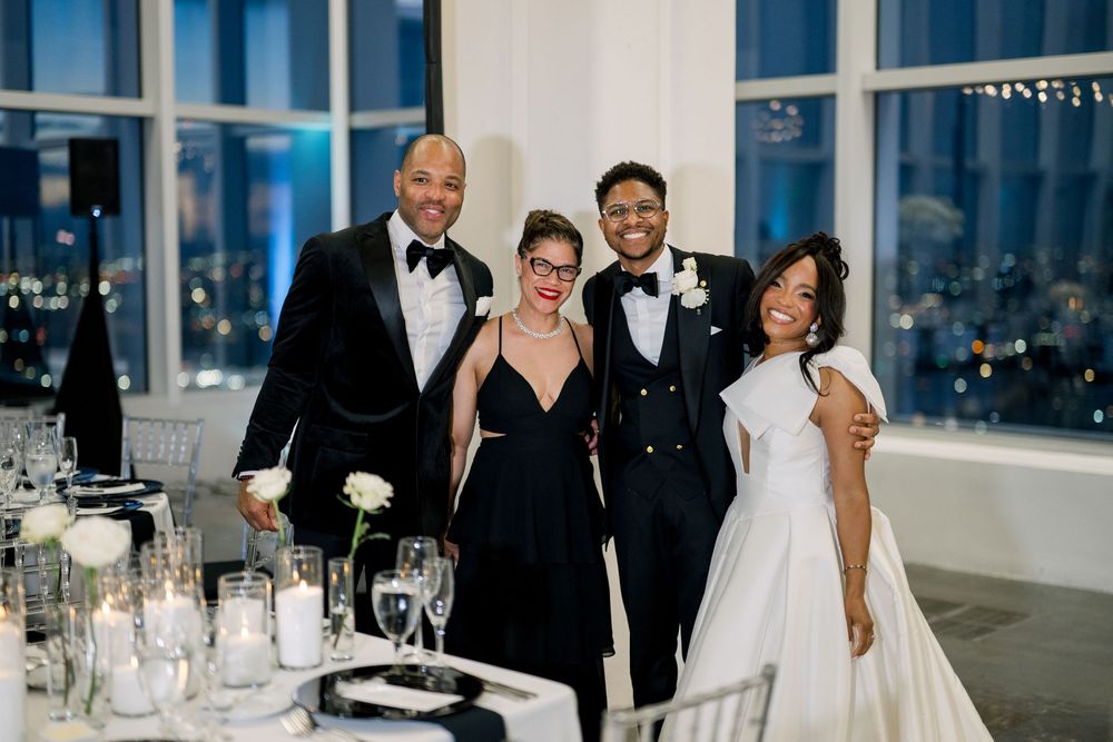 Wedding celebration group stands together in formal attire at an elegant reception venue with city views and candlelit tables.