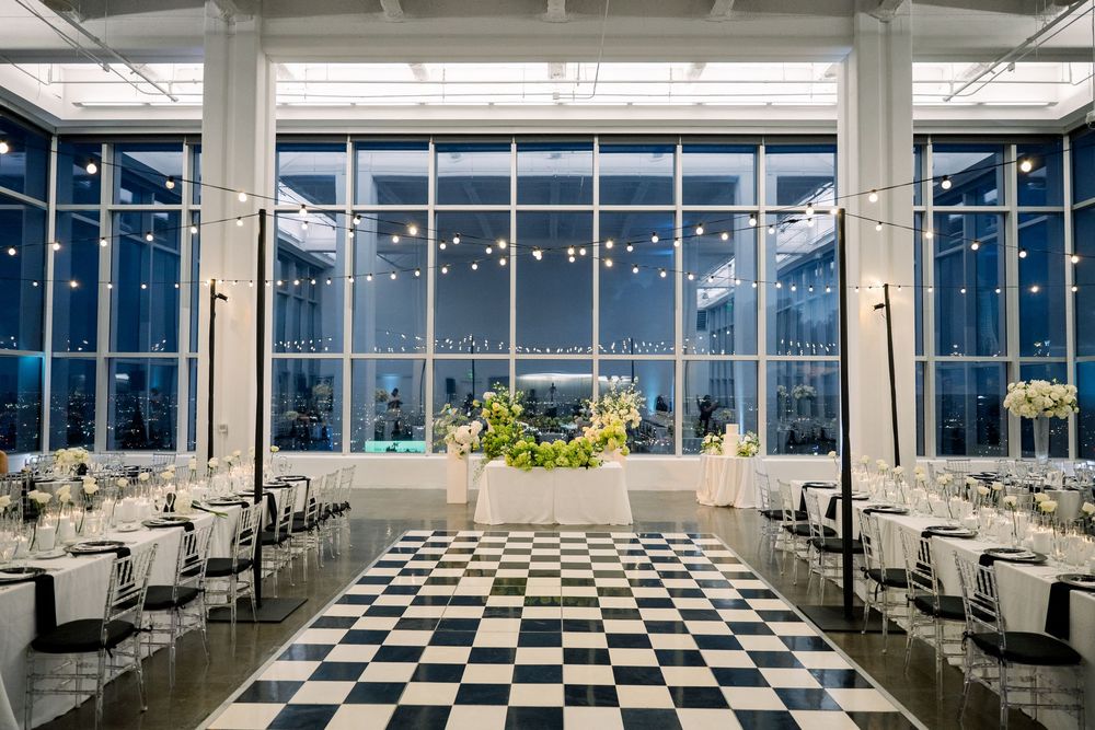 Modern event venue with floor-to-ceiling windows, checkered floor, and string lights overlooking a city skyline at dusk.